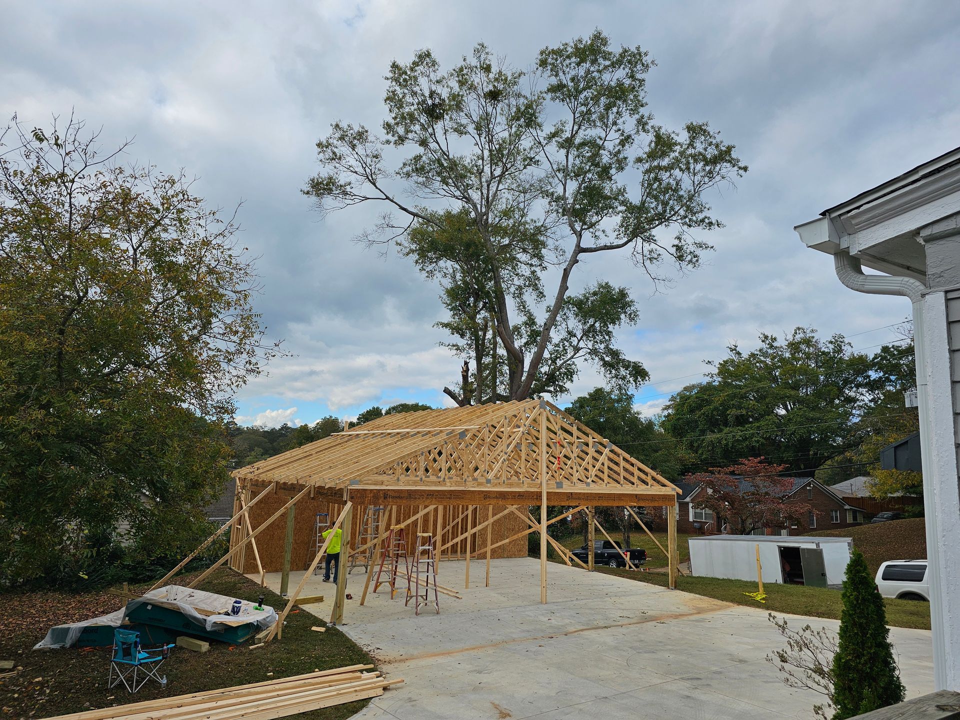 Construction of a wooden carport with roof trusses, trees in the background, cloudy sky.