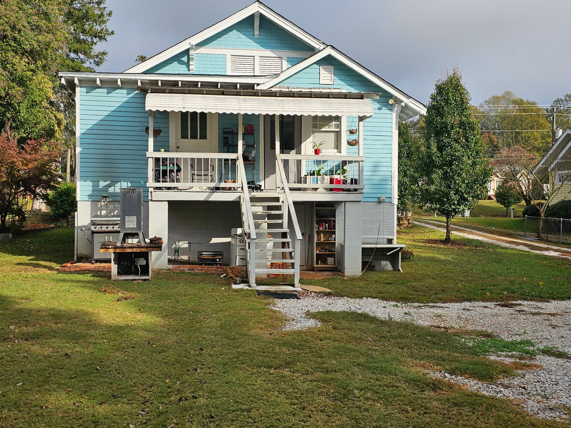 Blue house with a raised porch and wooden stairs. Gravel driveway, green grass, and trees.