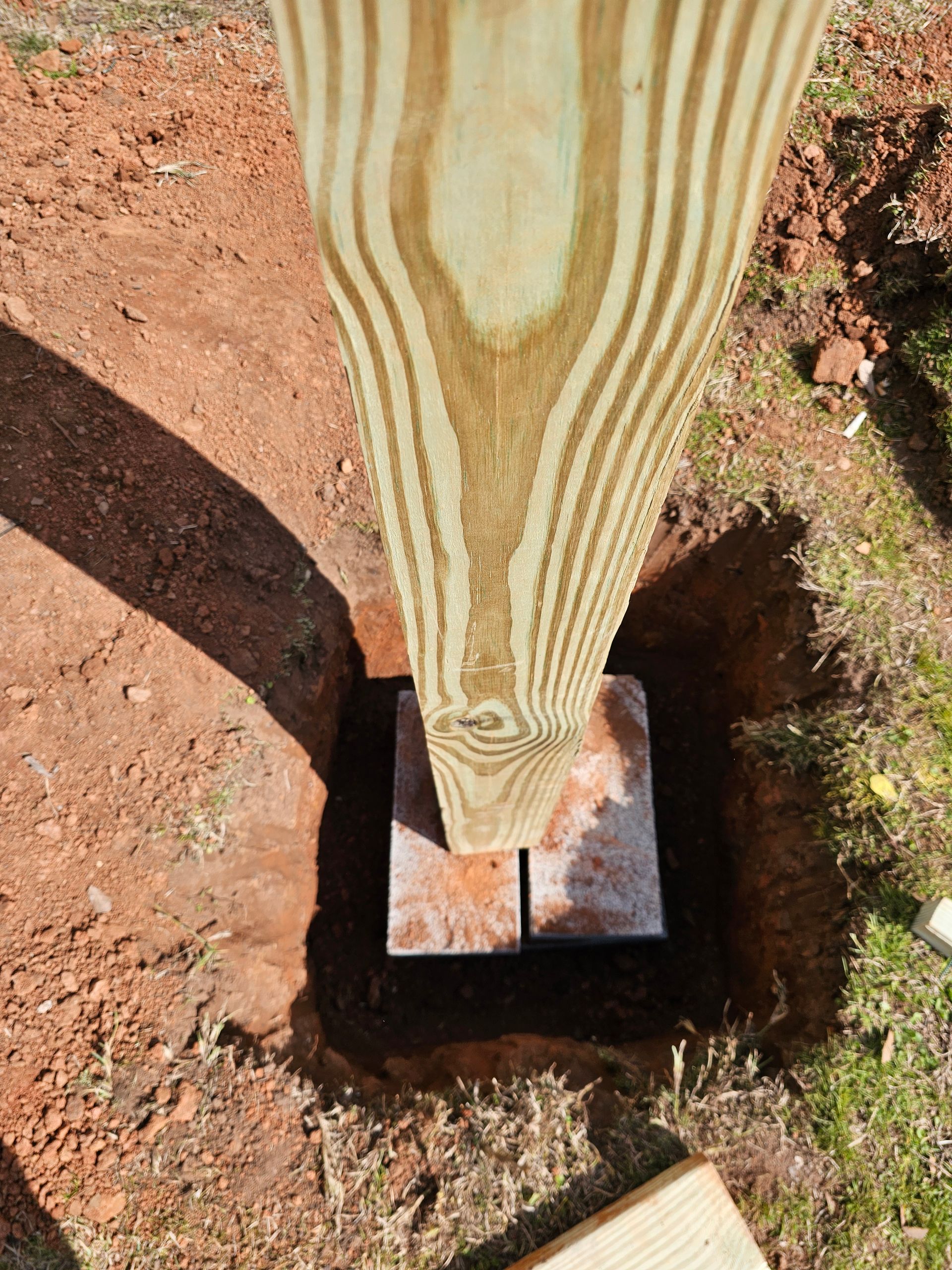 Wooden post being set in a square hole on top of two rectangular concrete blocks in dirt.
