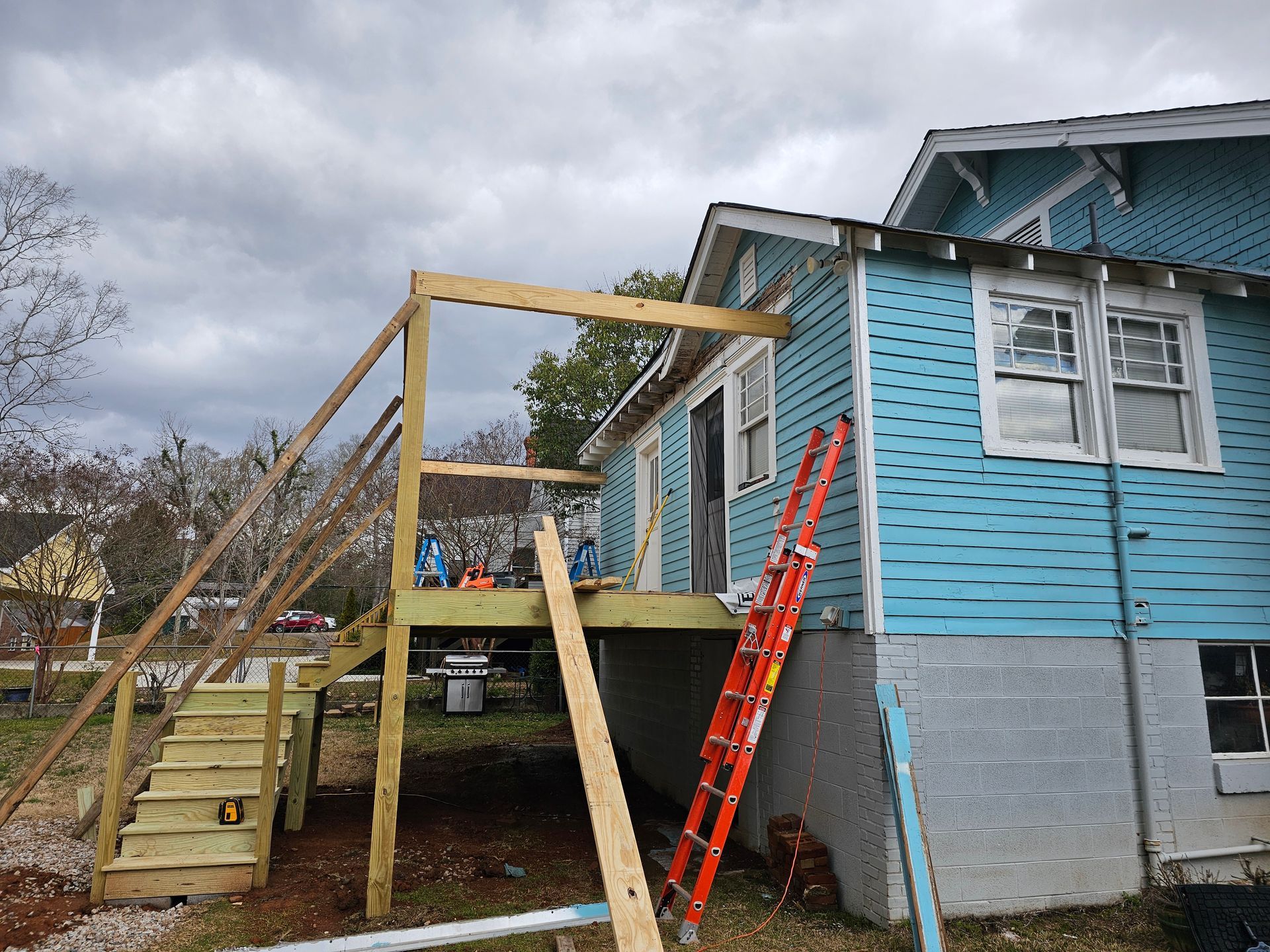 Wooden deck under construction next to a blue house with stairs and a ladder.