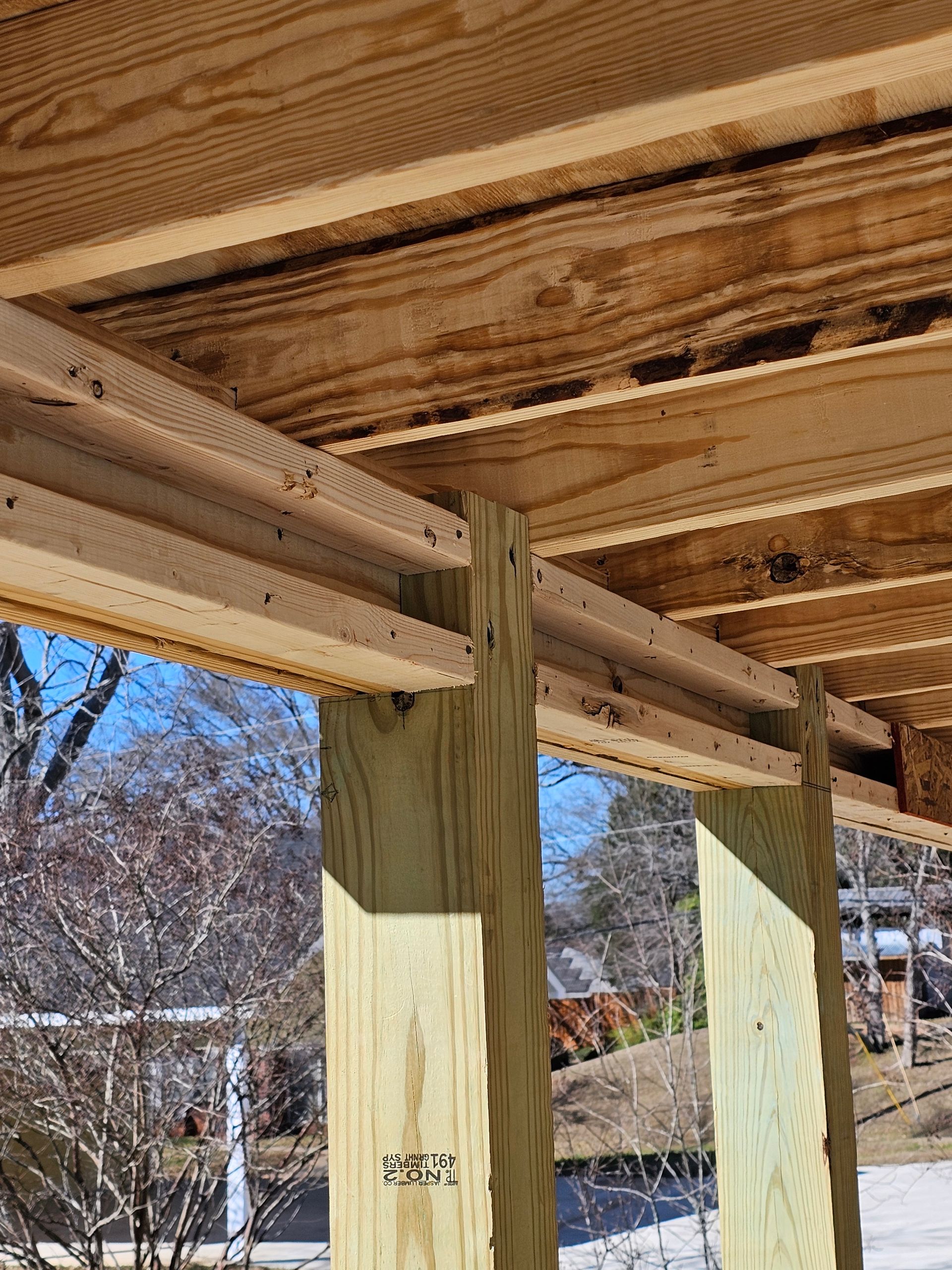 View from beneath a wooden deck, showing support beams, joists, and decking boards with sky and trees in background.