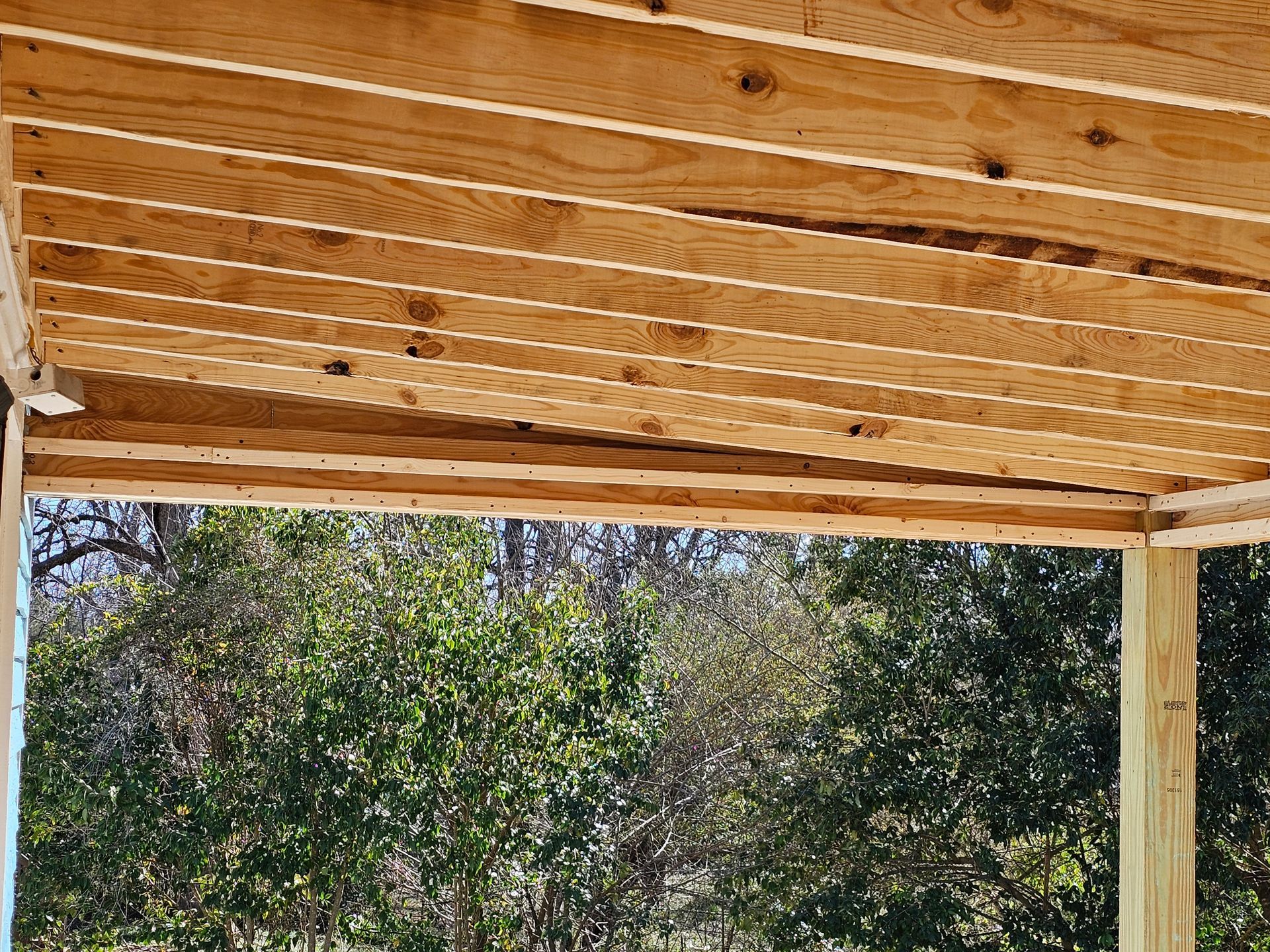Wooden porch ceiling with exposed beams, viewed against a backdrop of green trees.