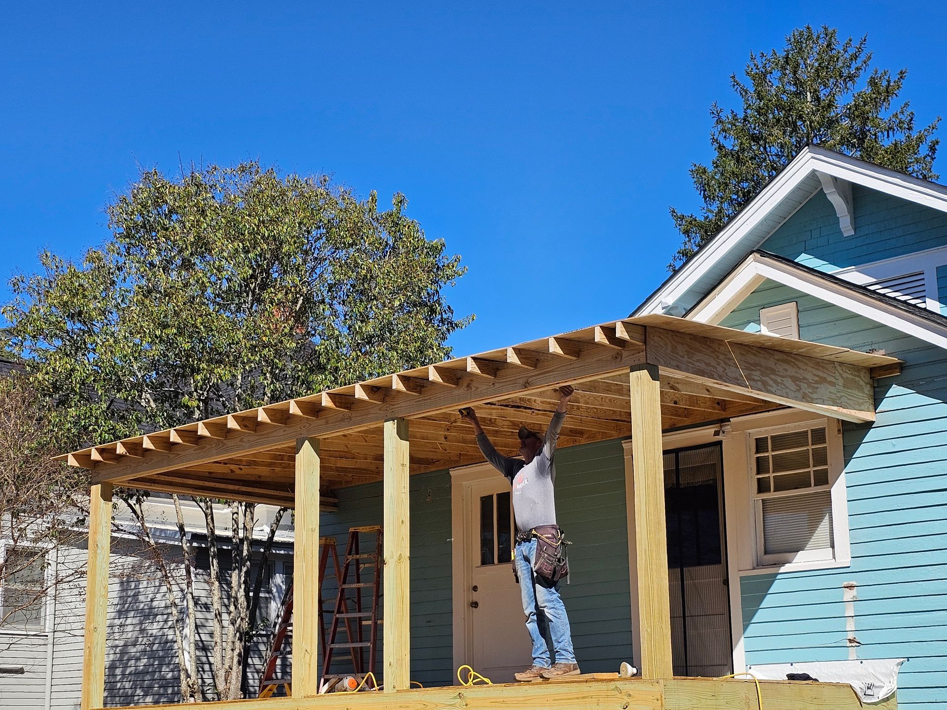 Construction worker building a wooden porch roof on a house; blue sky background.