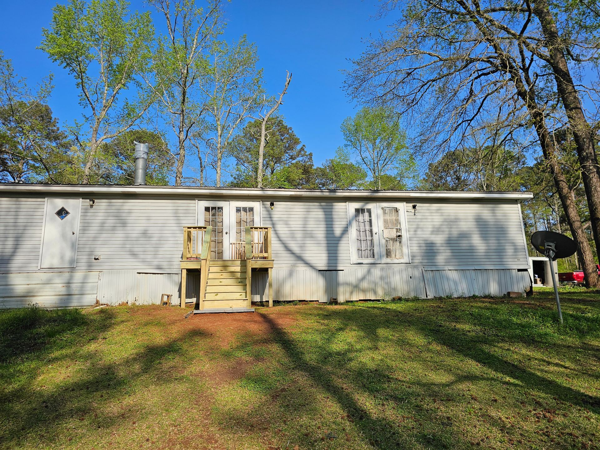 Mobile home exterior with wooden steps, surrounded by trees and grass, under a blue sky.