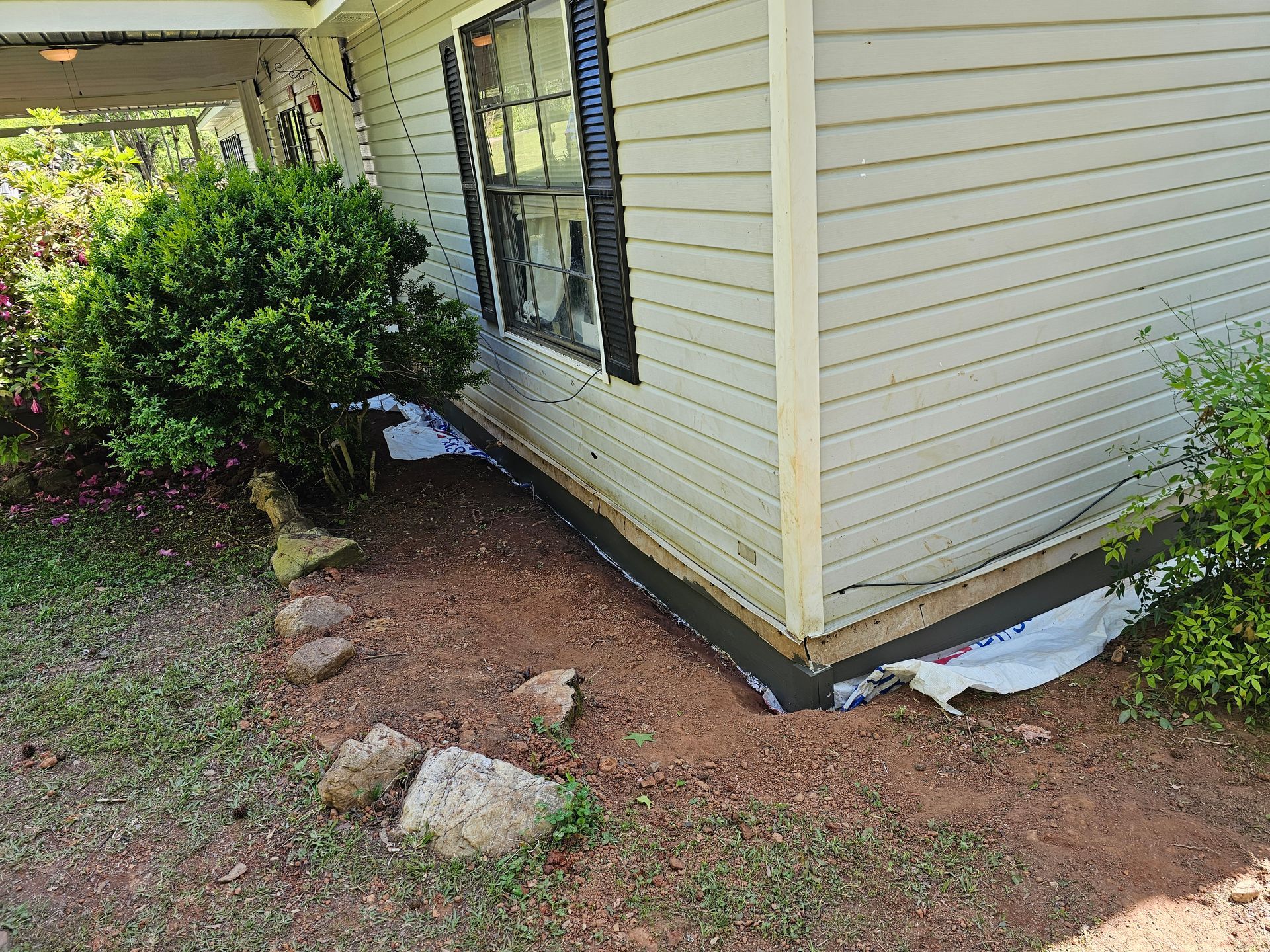 Side of a house with light siding and dark trim, exposed dirt, and landscaping.