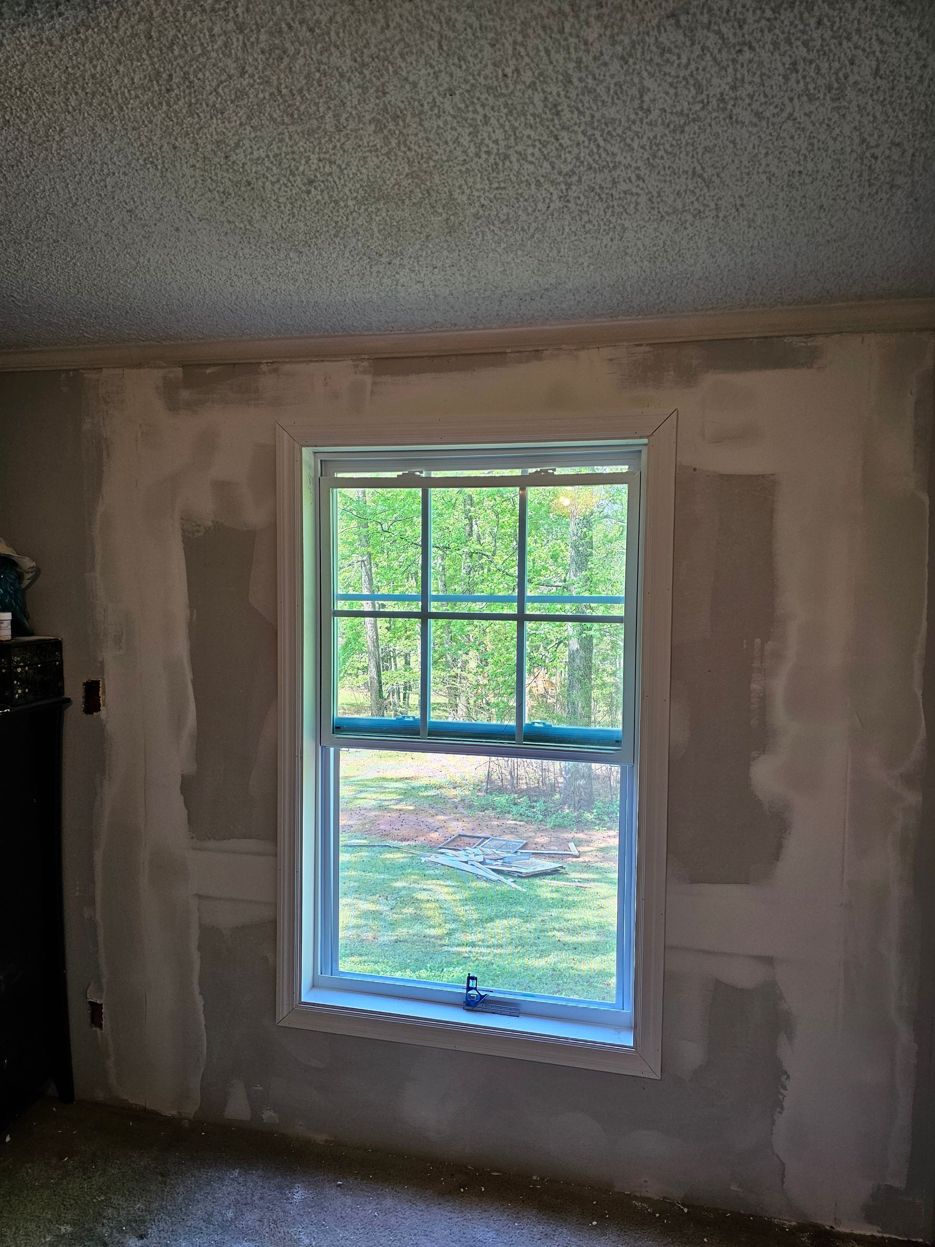 Window in a room with unfinished drywall; view of trees outside.
