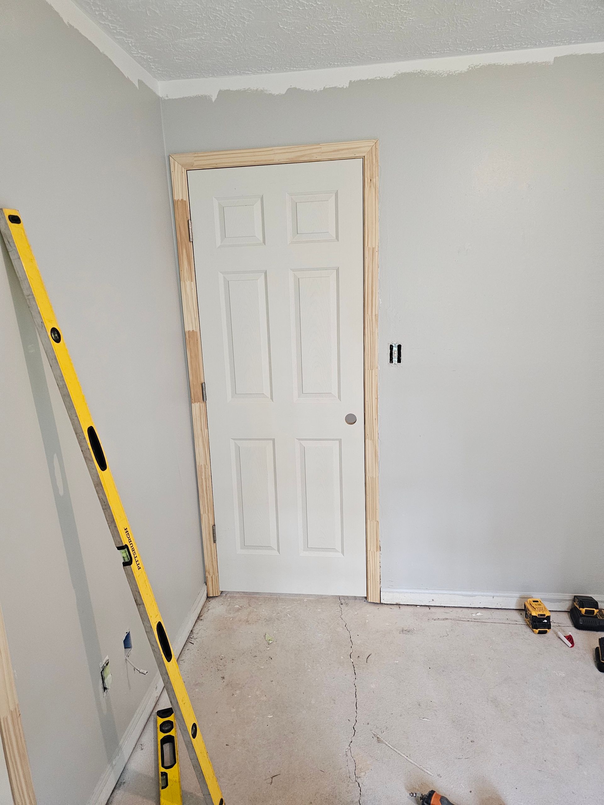Interior view of a door frame with trim being installed. A yellow level leans against a wall in a room under construction.