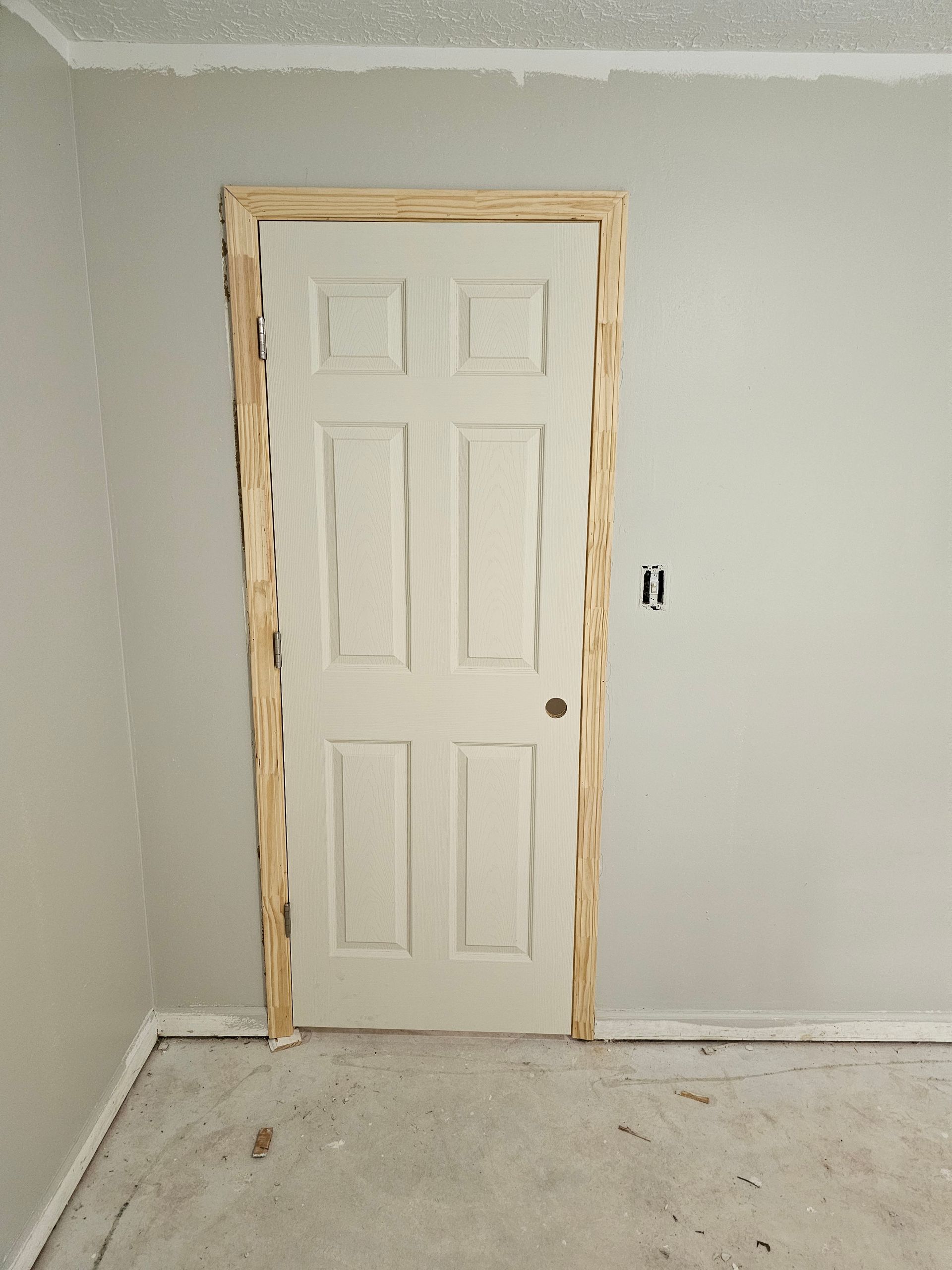 White six-panel door with unfinished wood trim in a room with gray walls and a concrete floor.