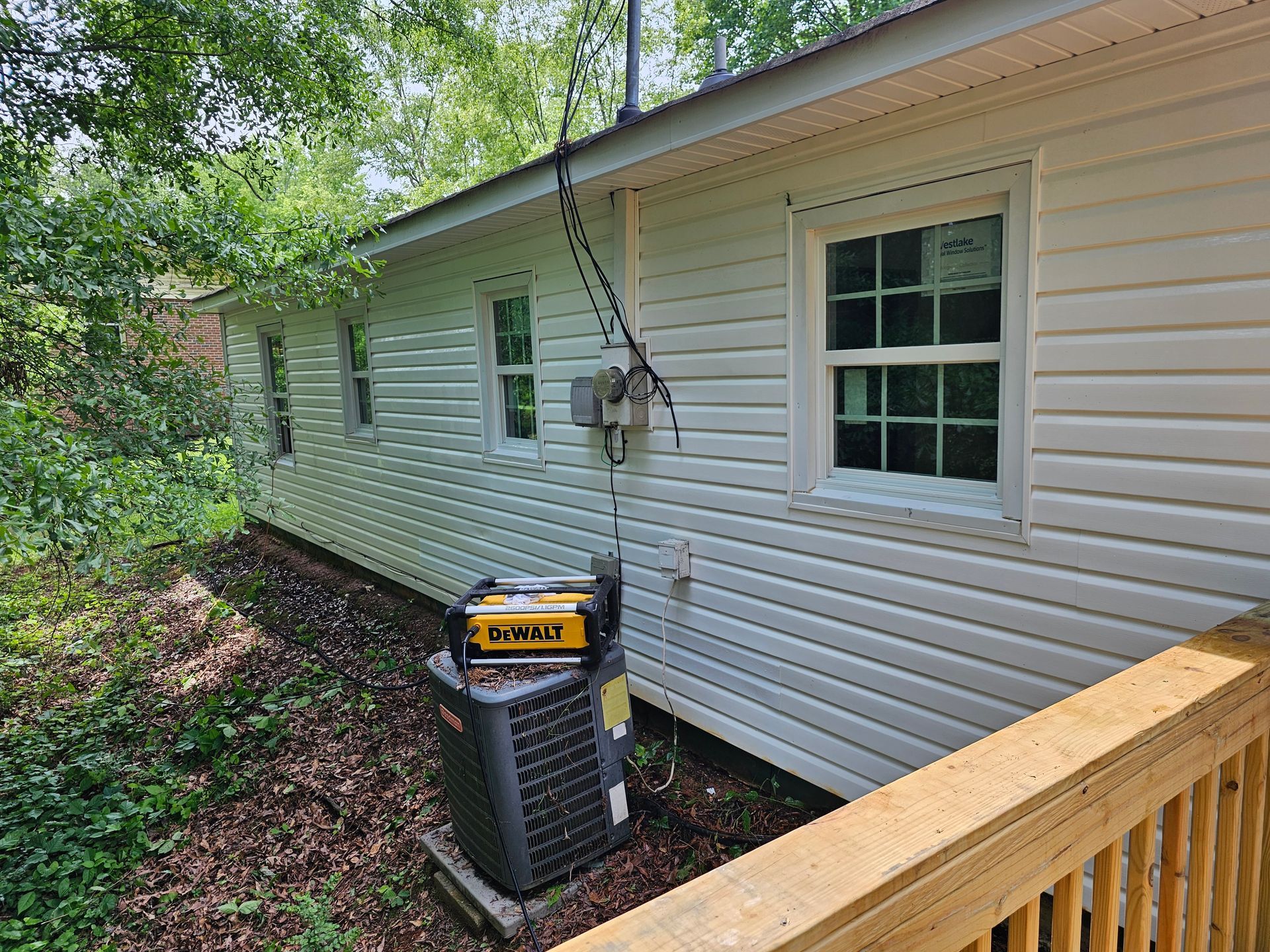 Side of a white house with windows, air conditioner, and generator, with trees in the background.