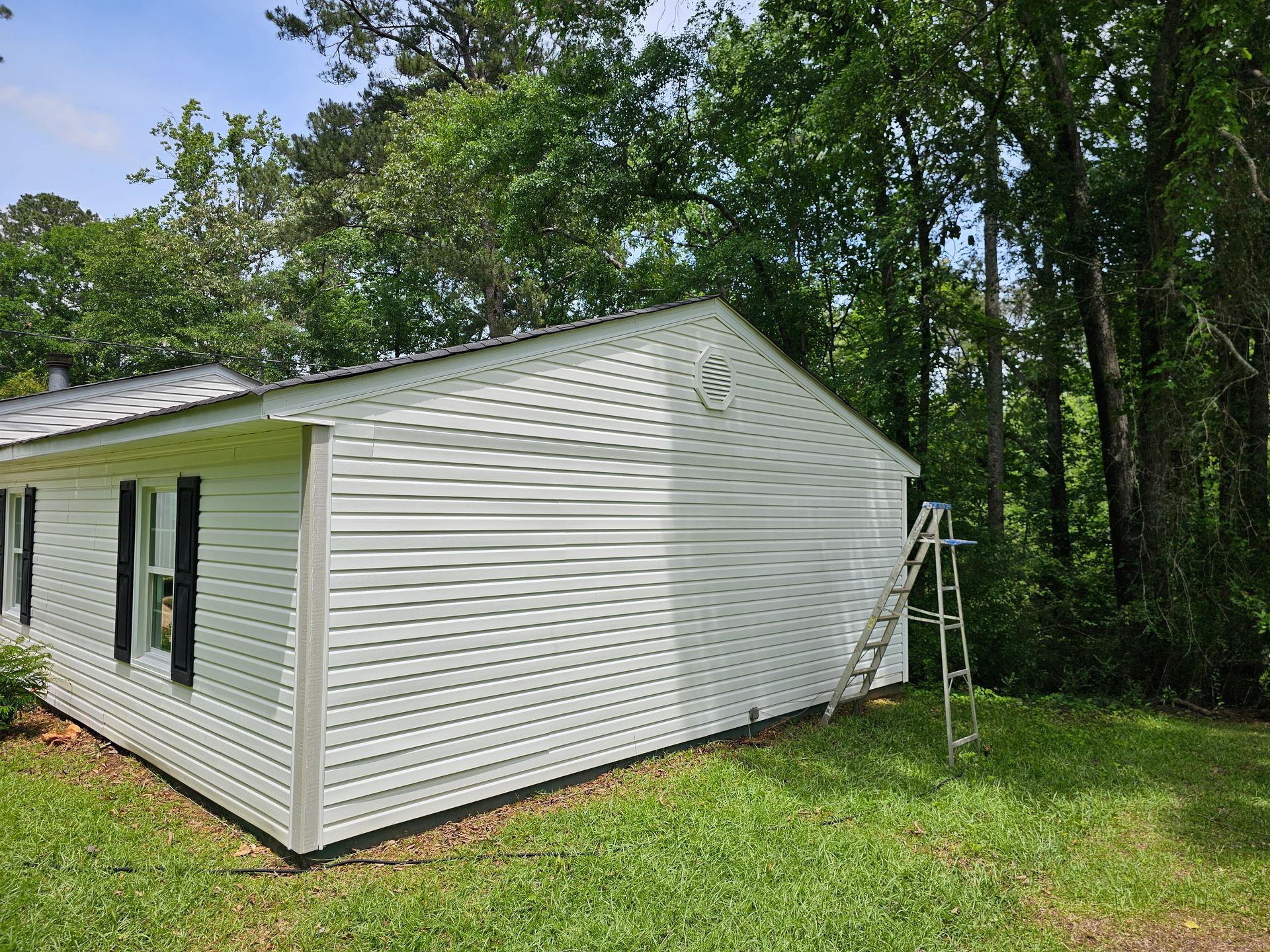 White-sided house with black shutters, ladder leaning against the side, surrounded by green trees and grass.