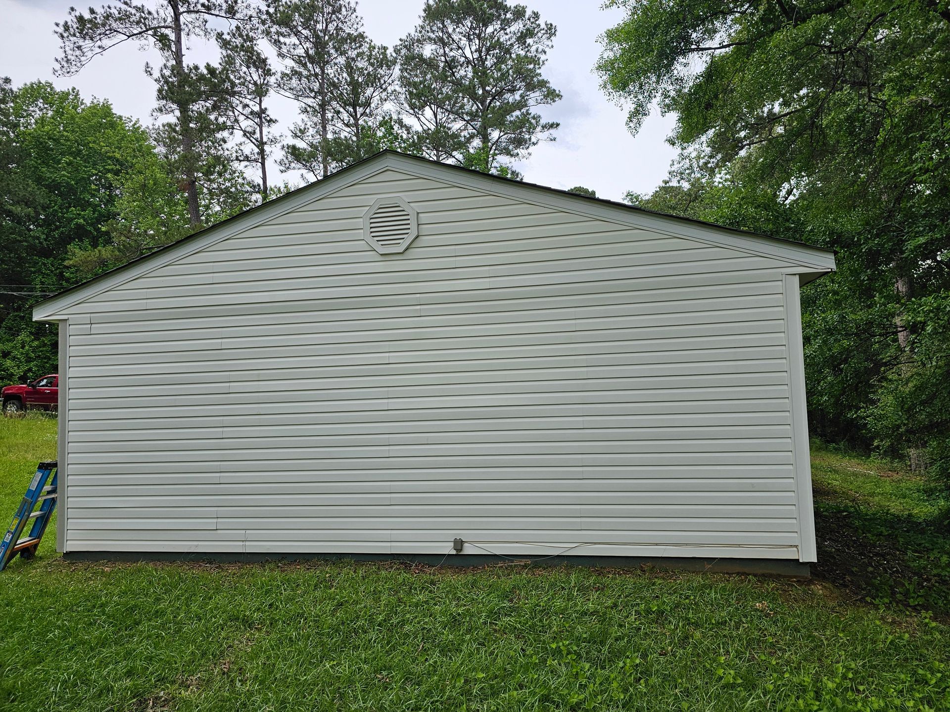 White-sided shed with dark roof and circular vent, surrounded by trees and grass, on a cloudy day.