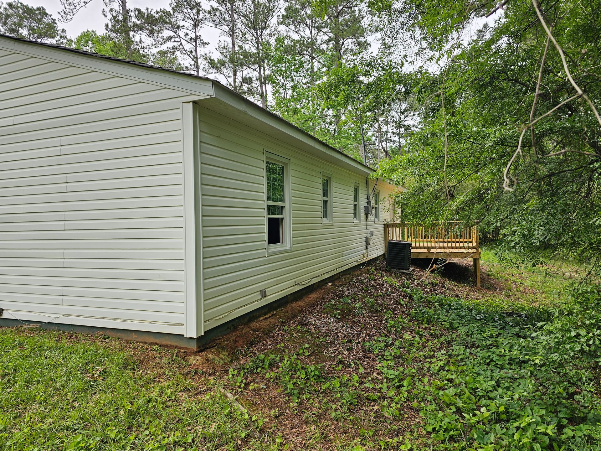 Side view of a light-colored house with a small wooden deck surrounded by trees and vegetation.
