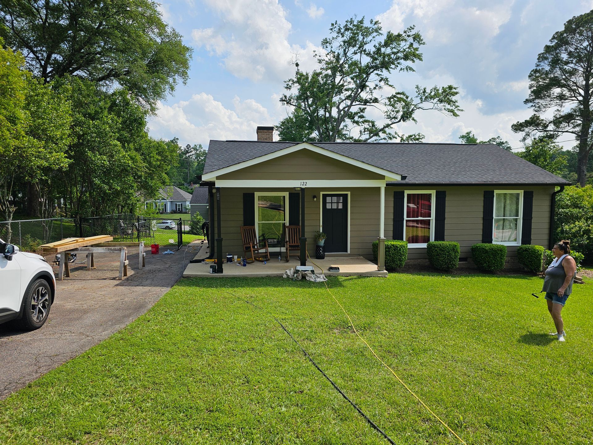 Tan house with black shutters, green lawn, gravel driveway, person standing on grass.