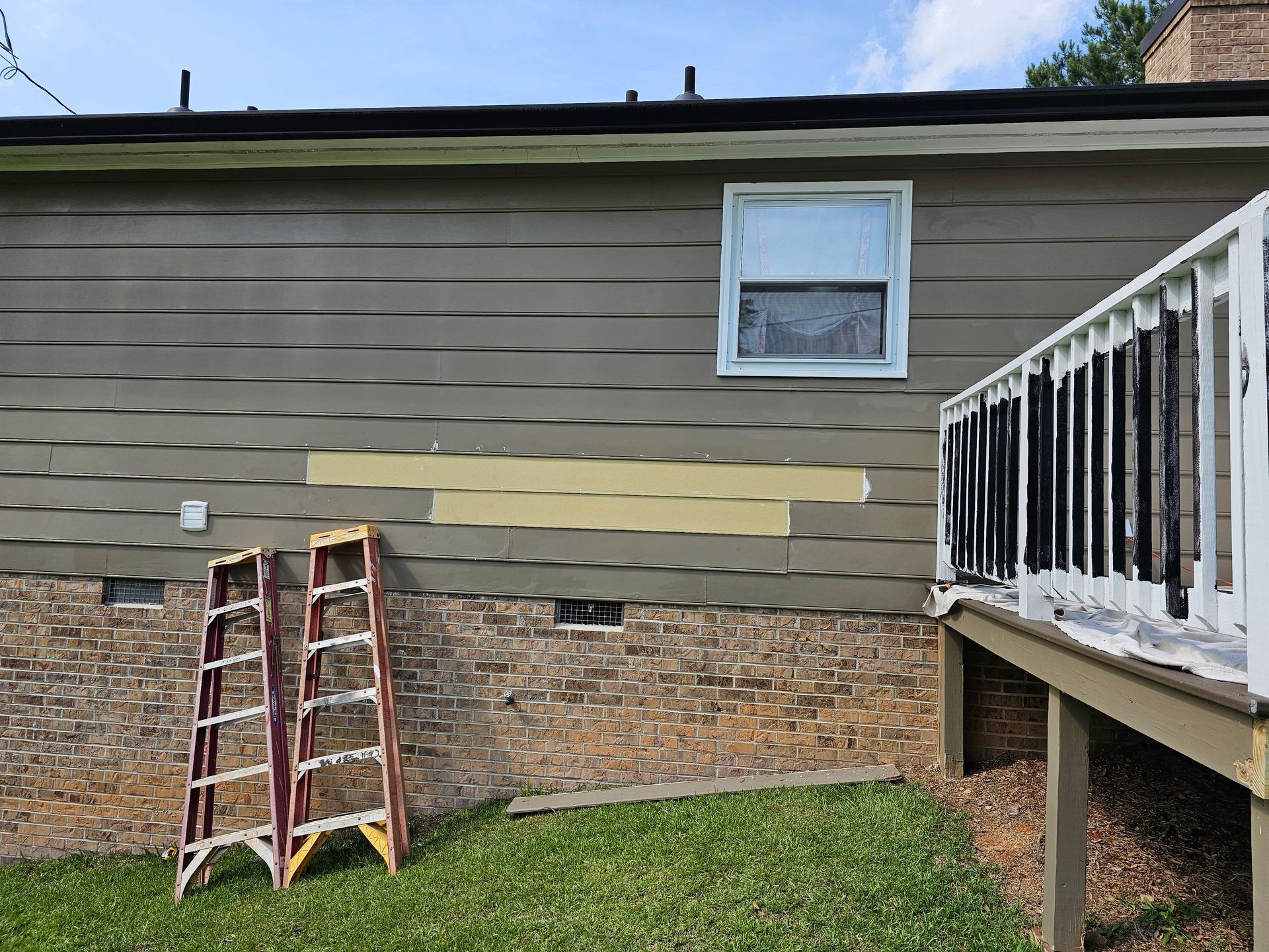 House exterior with brown siding, a window, and a deck. Two ladders lean against the wall.