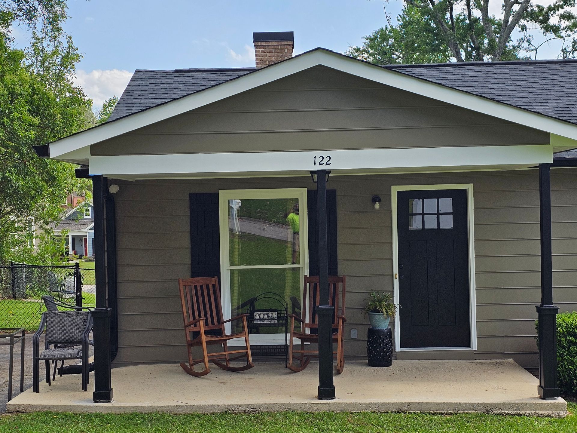 Tan house with black trim and door, porch with rocking chairs and a concrete foundation.