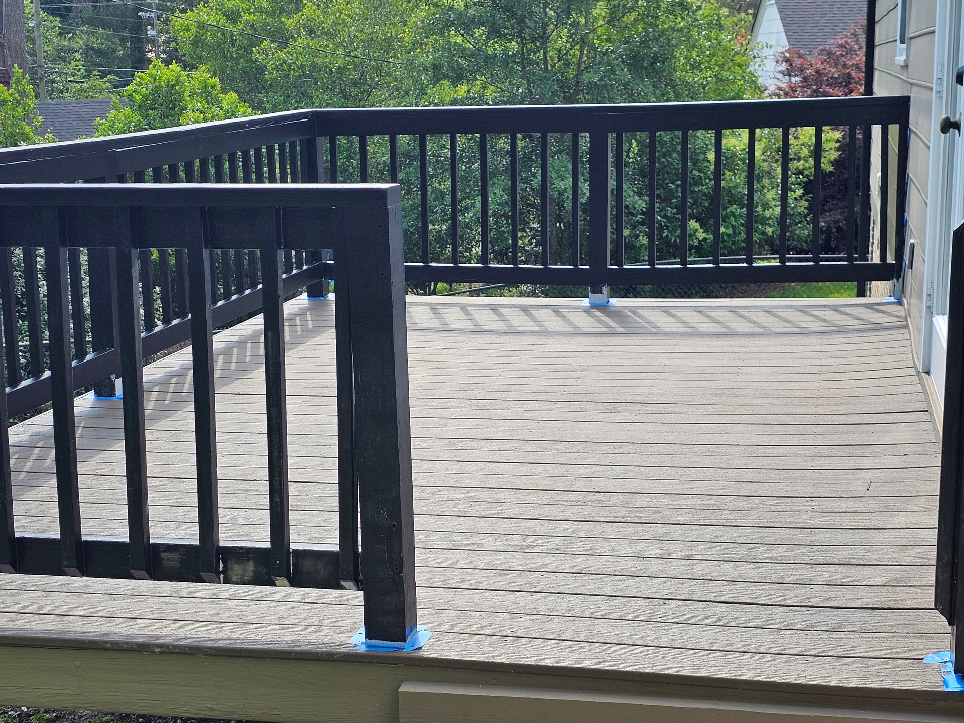 Black railed deck with gray textured surface. Trees and house visible in background.