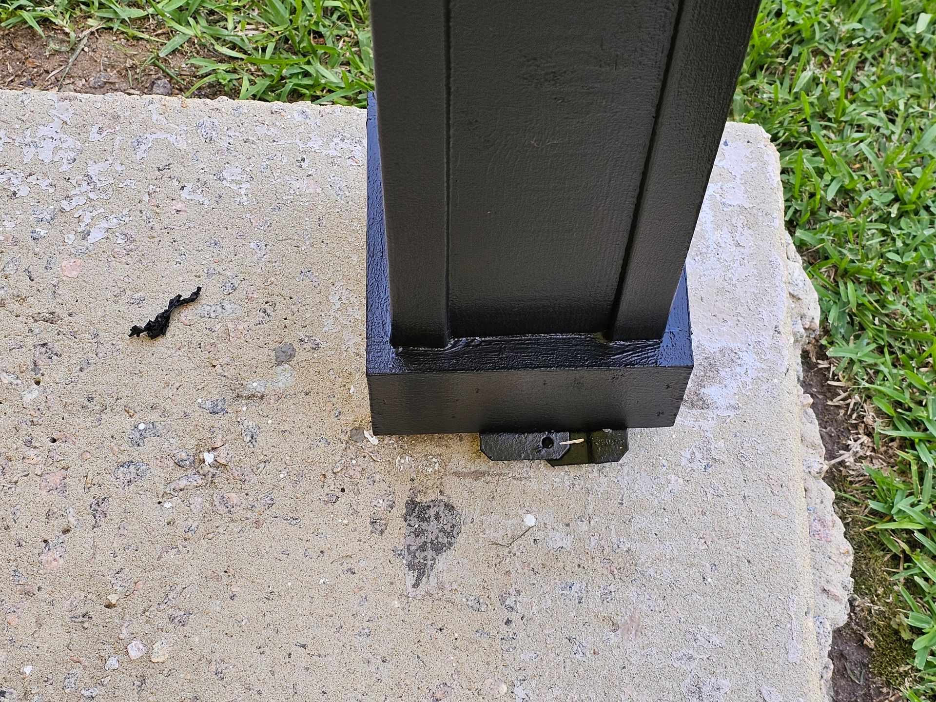 Black post attached to concrete base with bolt. Green grass in background.