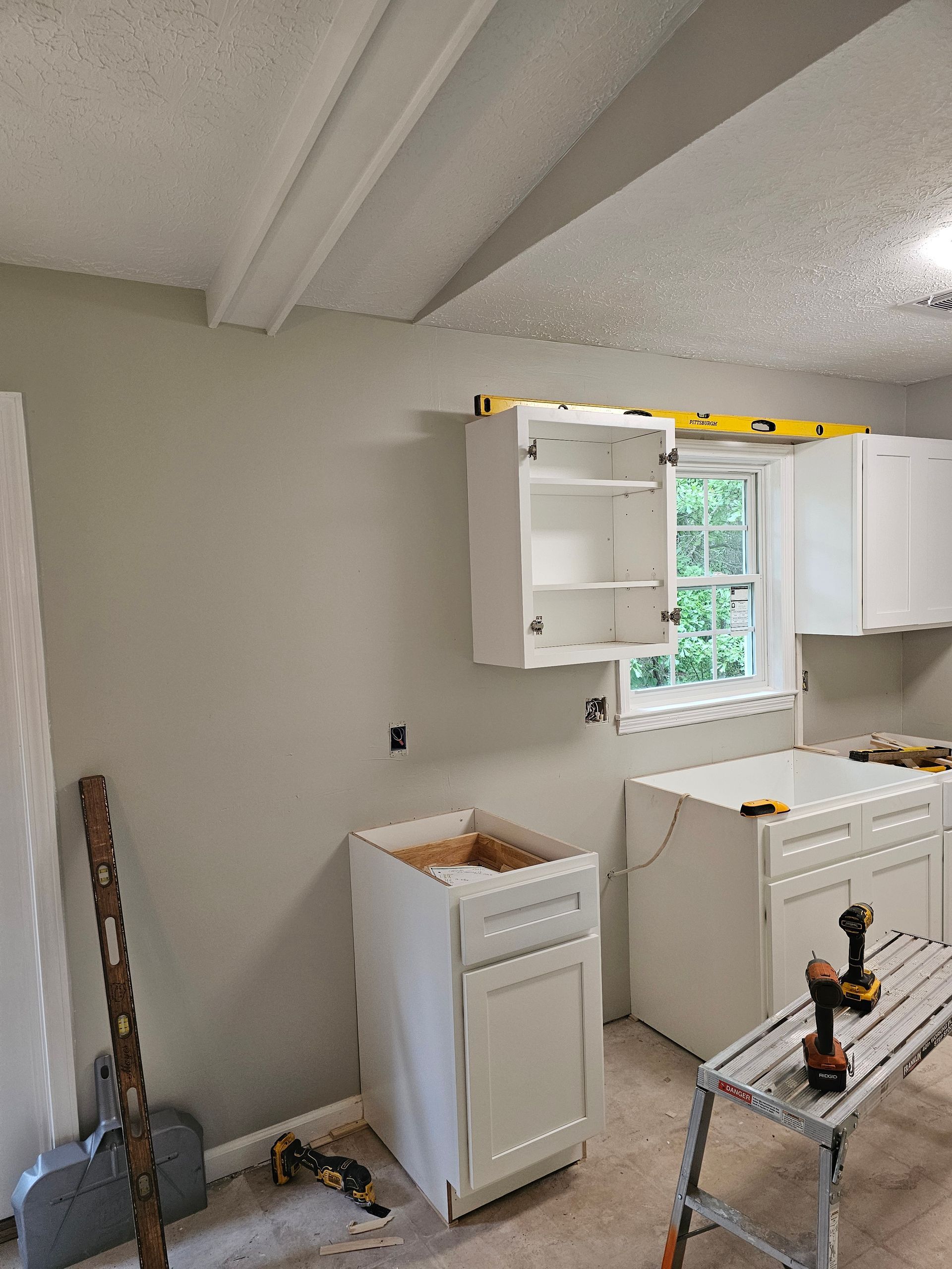 Kitchen cabinets being installed; white cabinets, level on upper cabinet, beige walls, exposed ceiling beams, unfinished.