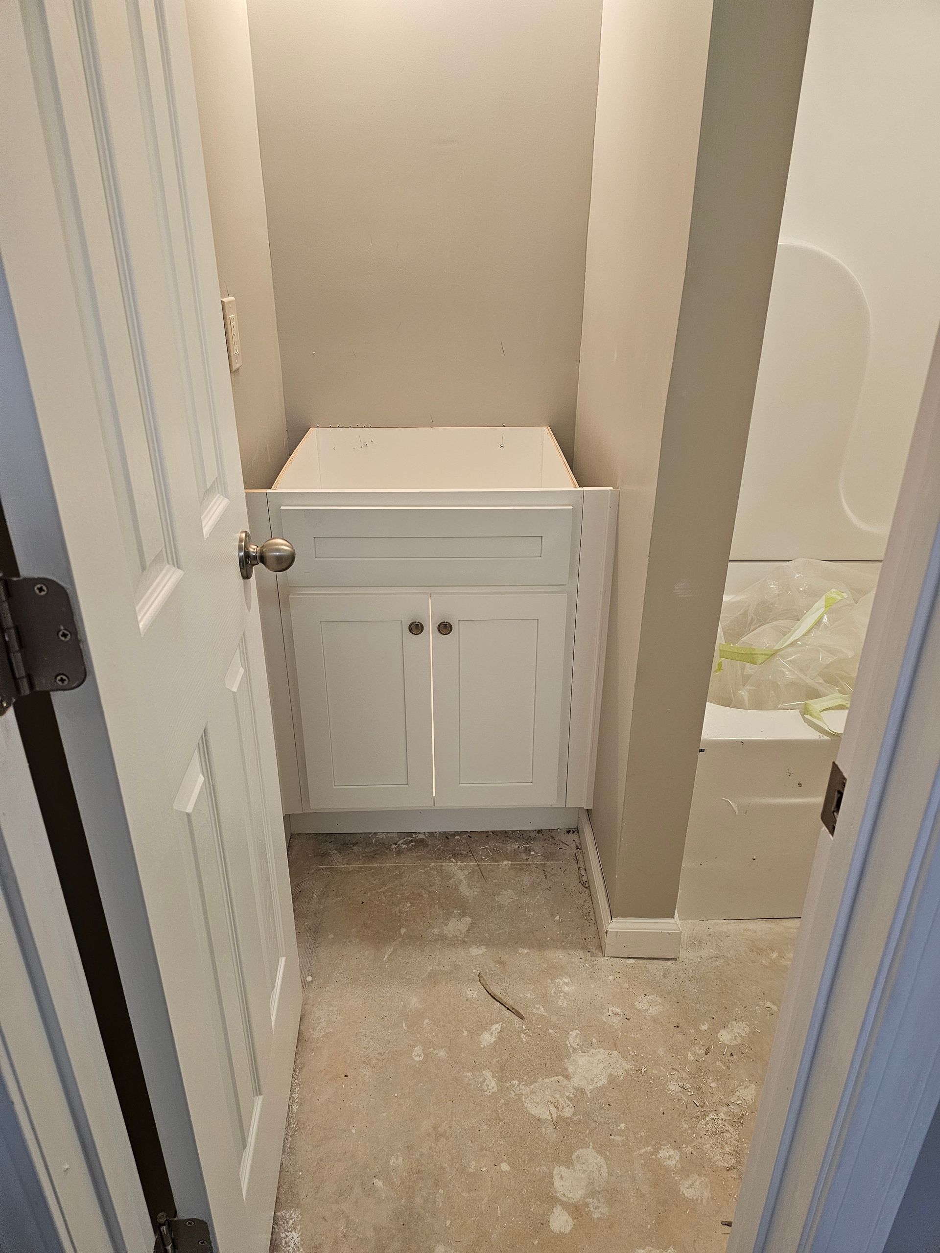 White bathroom vanity between walls, unfinished floor. Door on left, tub on right.