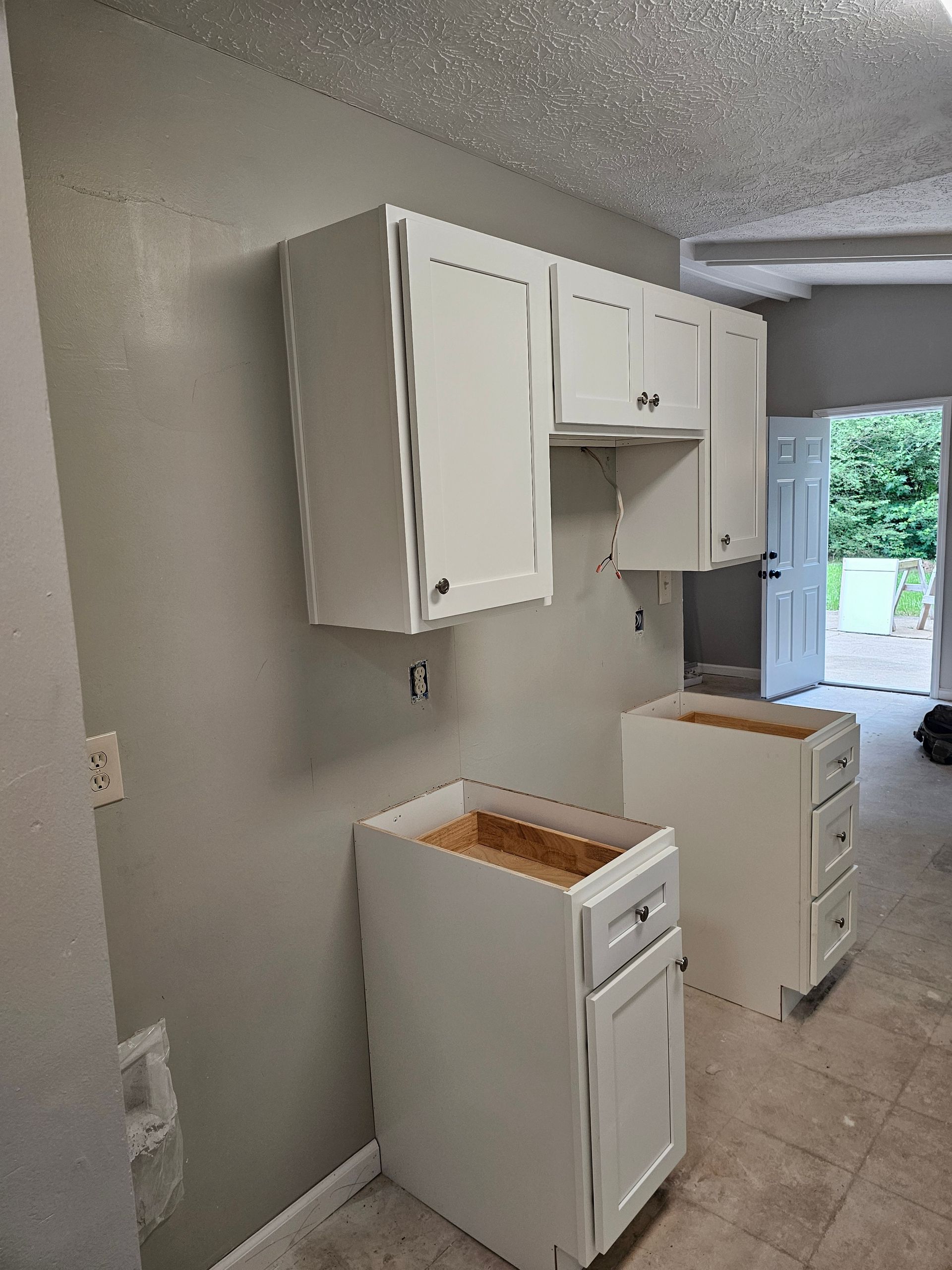 White kitchen cabinets mounted on a gray wall in a room under construction.