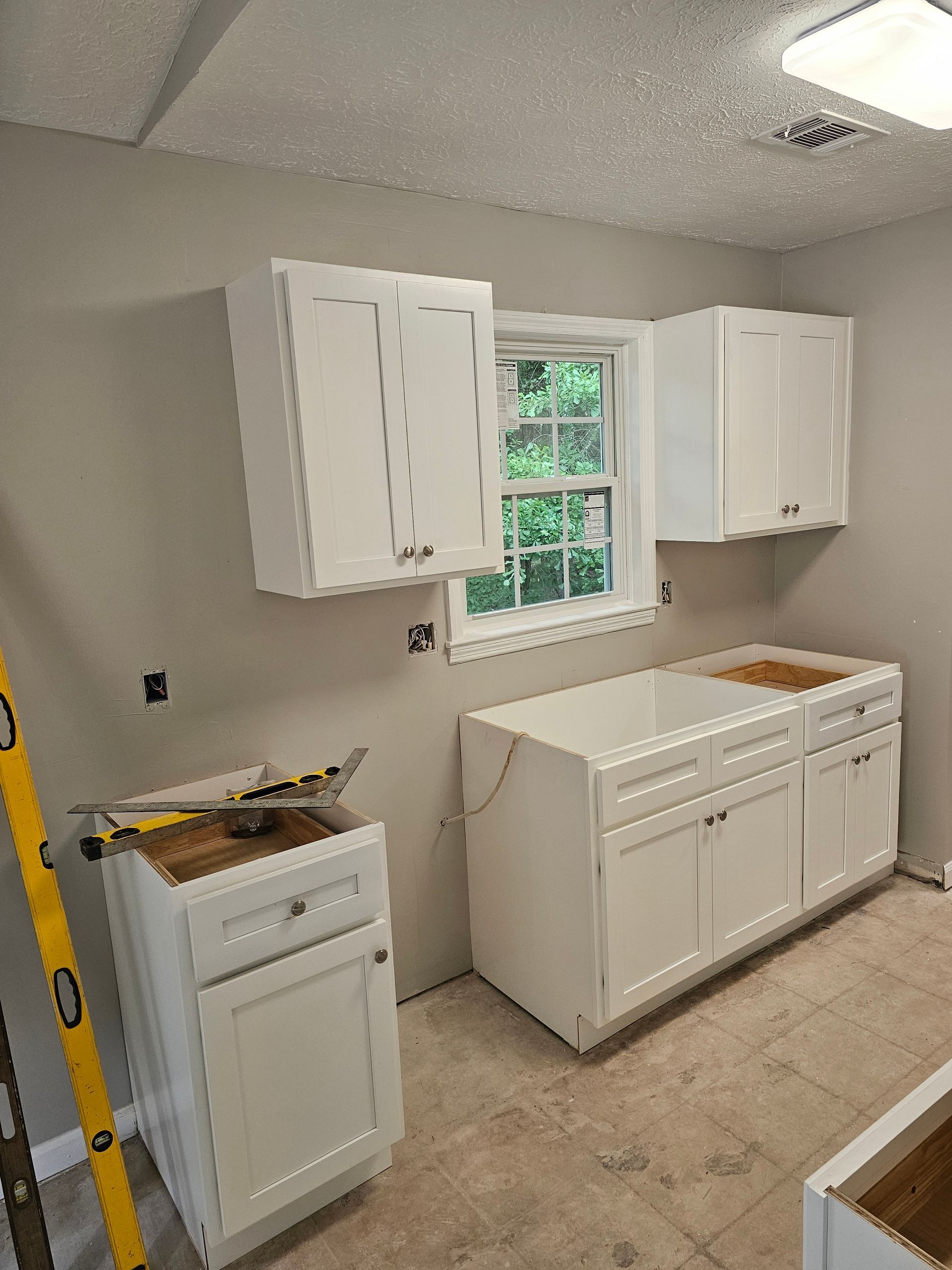 White kitchen cabinets being installed in a room with a window, beige walls, and tan flooring.