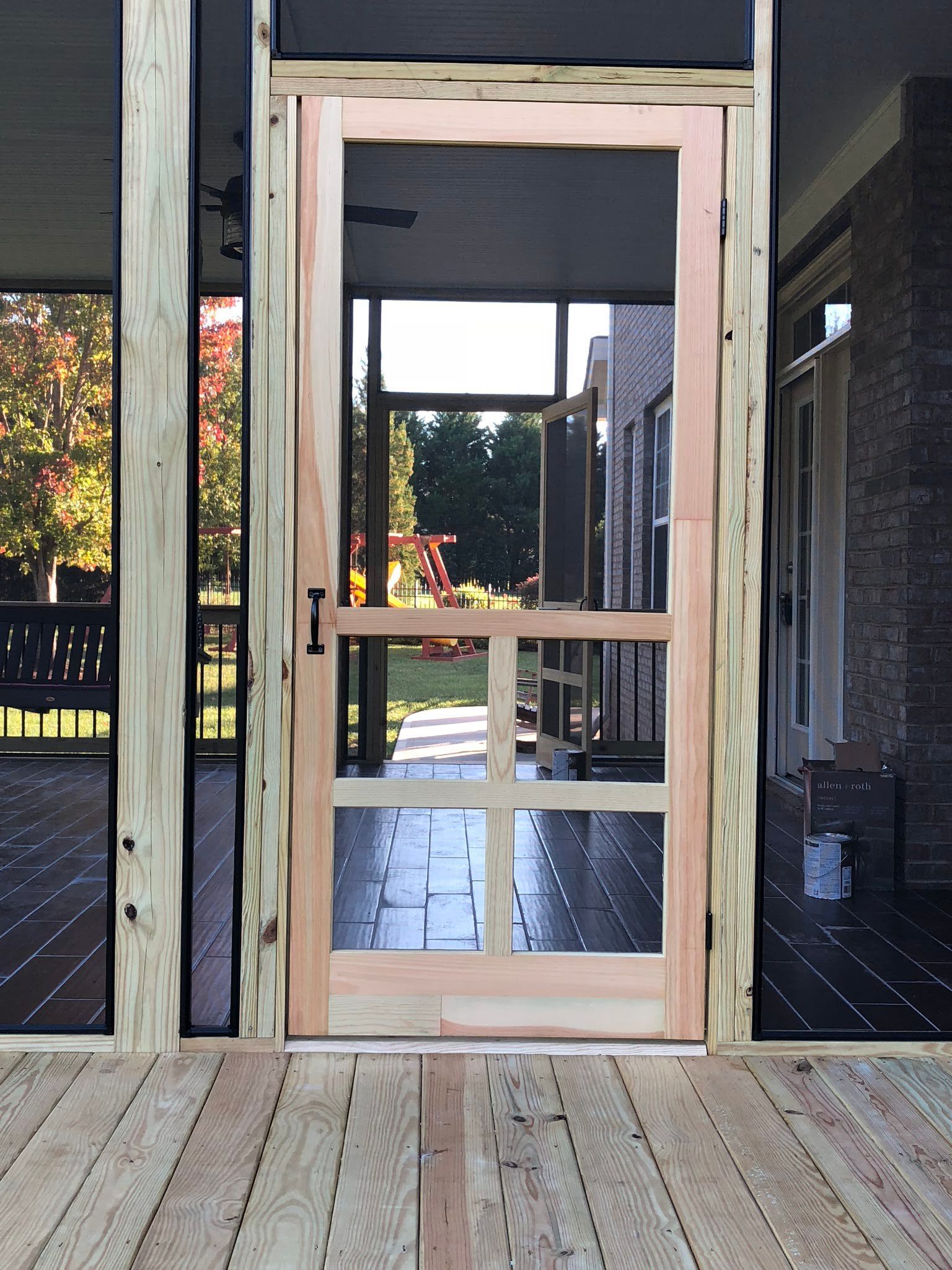 Screen door on a porch, view to the backyard. Wooden door frame and porch, trees in the background.
