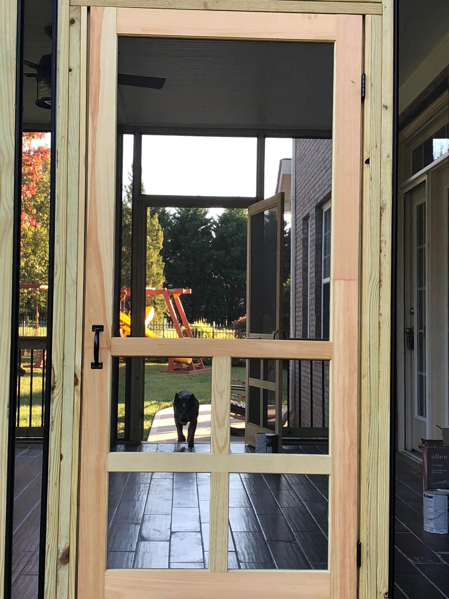 Screen door on a porch, view of a yard. Dog walks in, sunshine, brick house, and trees are visible.