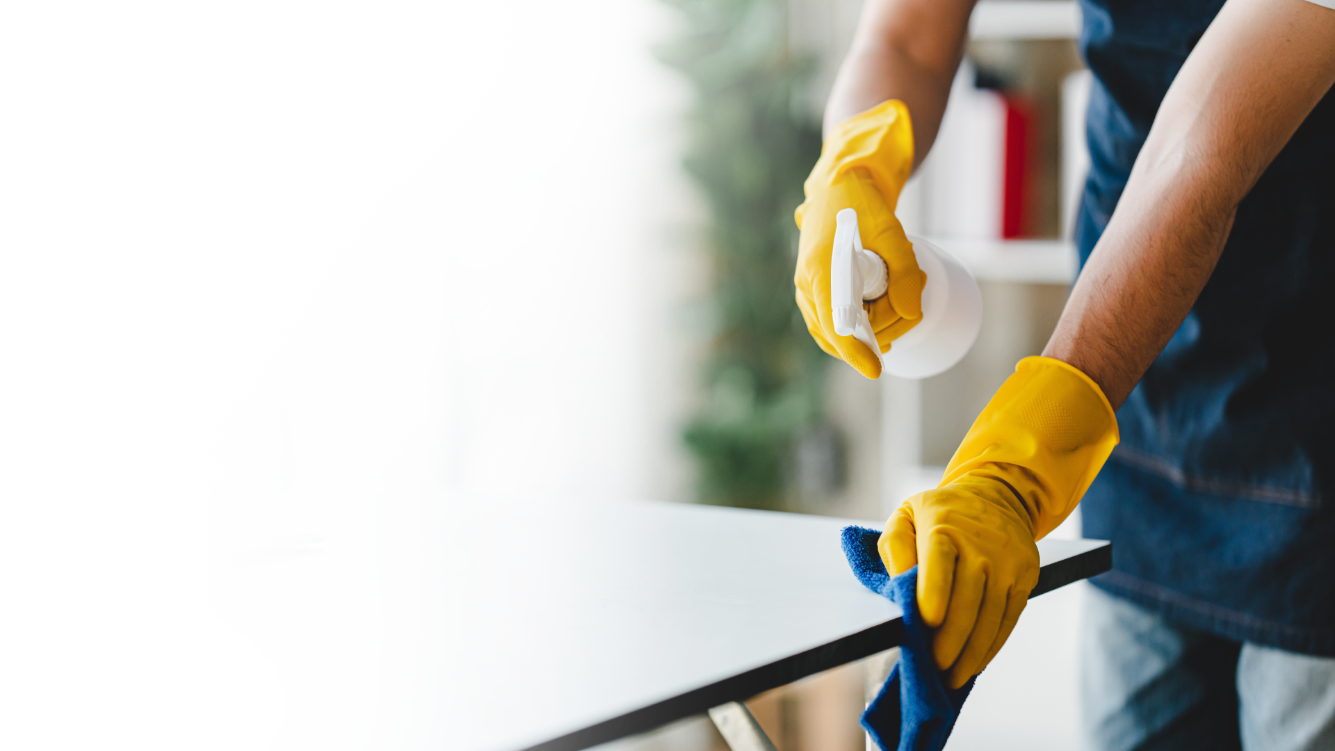 Person in yellow gloves spraying and wiping a surface with a blue cloth in a room.