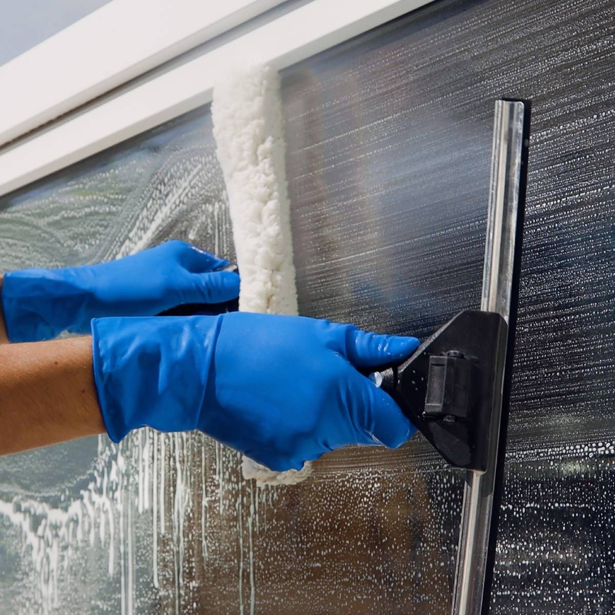 Hands in blue gloves cleaning a window with a soapy applicator and squeegee.