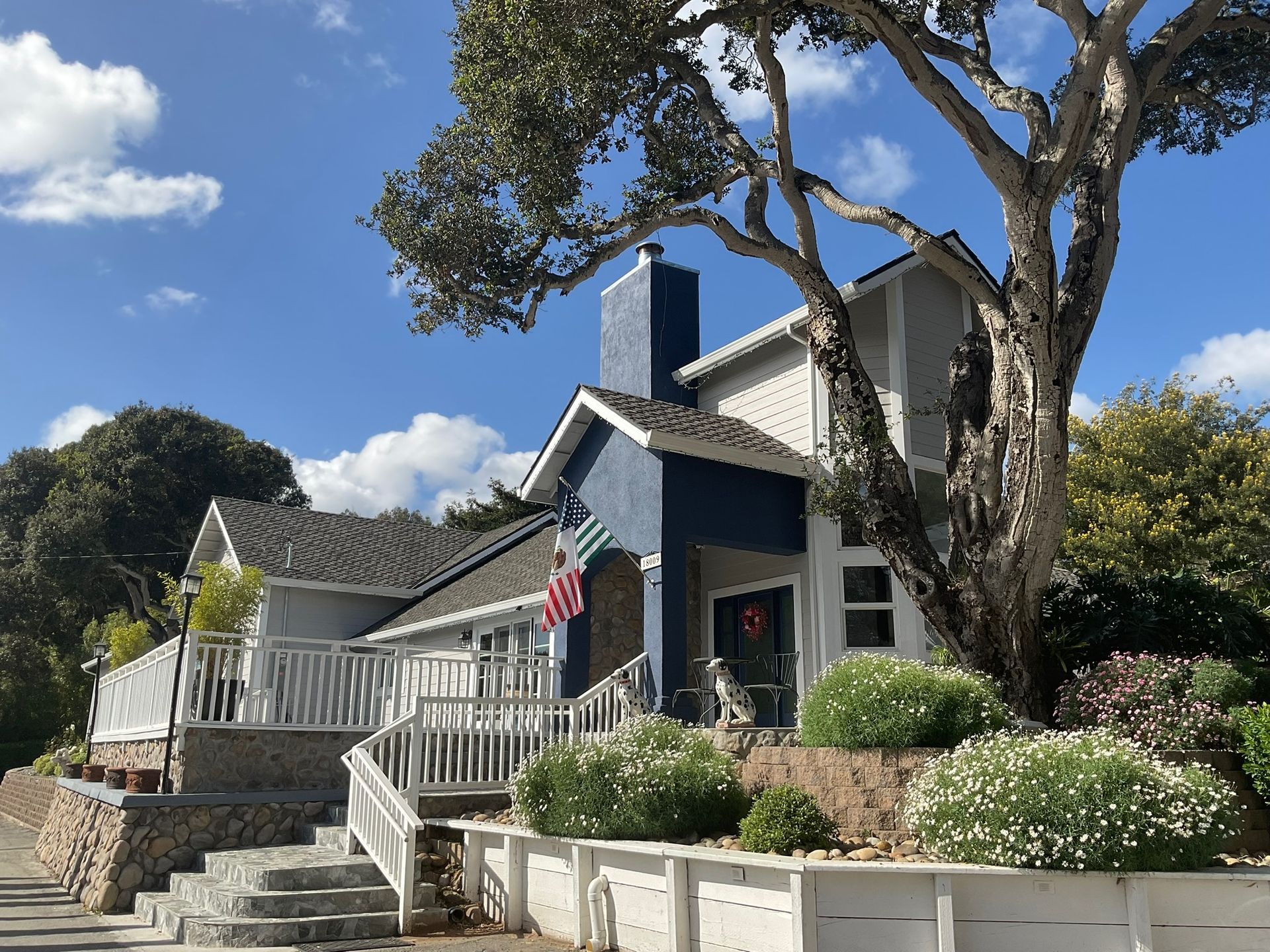 House with blue and gray exterior, stairs, white picket fence, large tree, and landscaping on a sunny day.