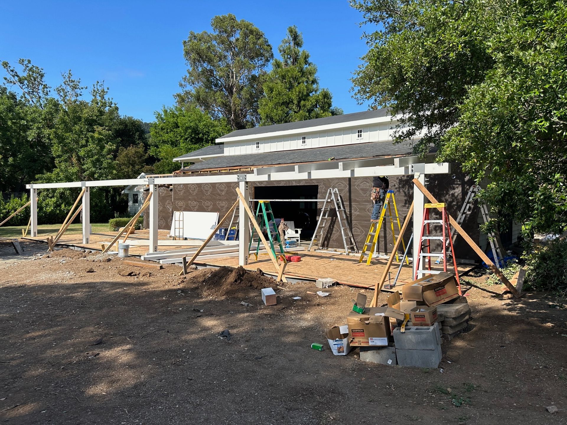 Construction site: white pergola being built next to a brown brick building. Tools and supports visible. Sunny day.