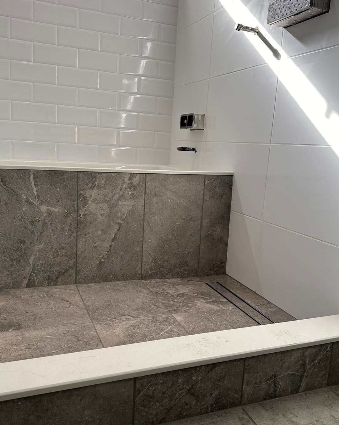 Bathroom with a tiled tub and shower area, featuring gray and white tiles, with sunlight.