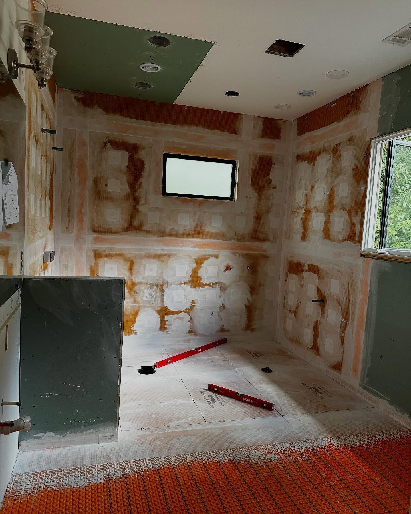 Bathroom under construction, orange walls, drywall removed, red level tools, open ceiling lights, window.
