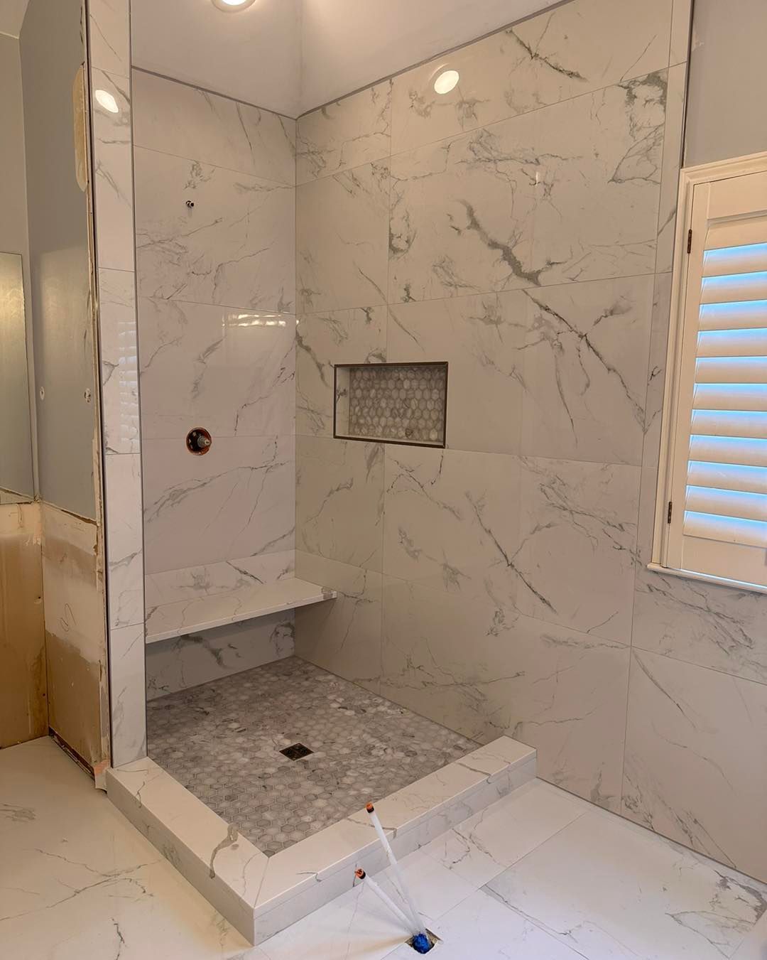 White marble tiled shower with built-in bench and niche, and a gray stone floor, next to window.