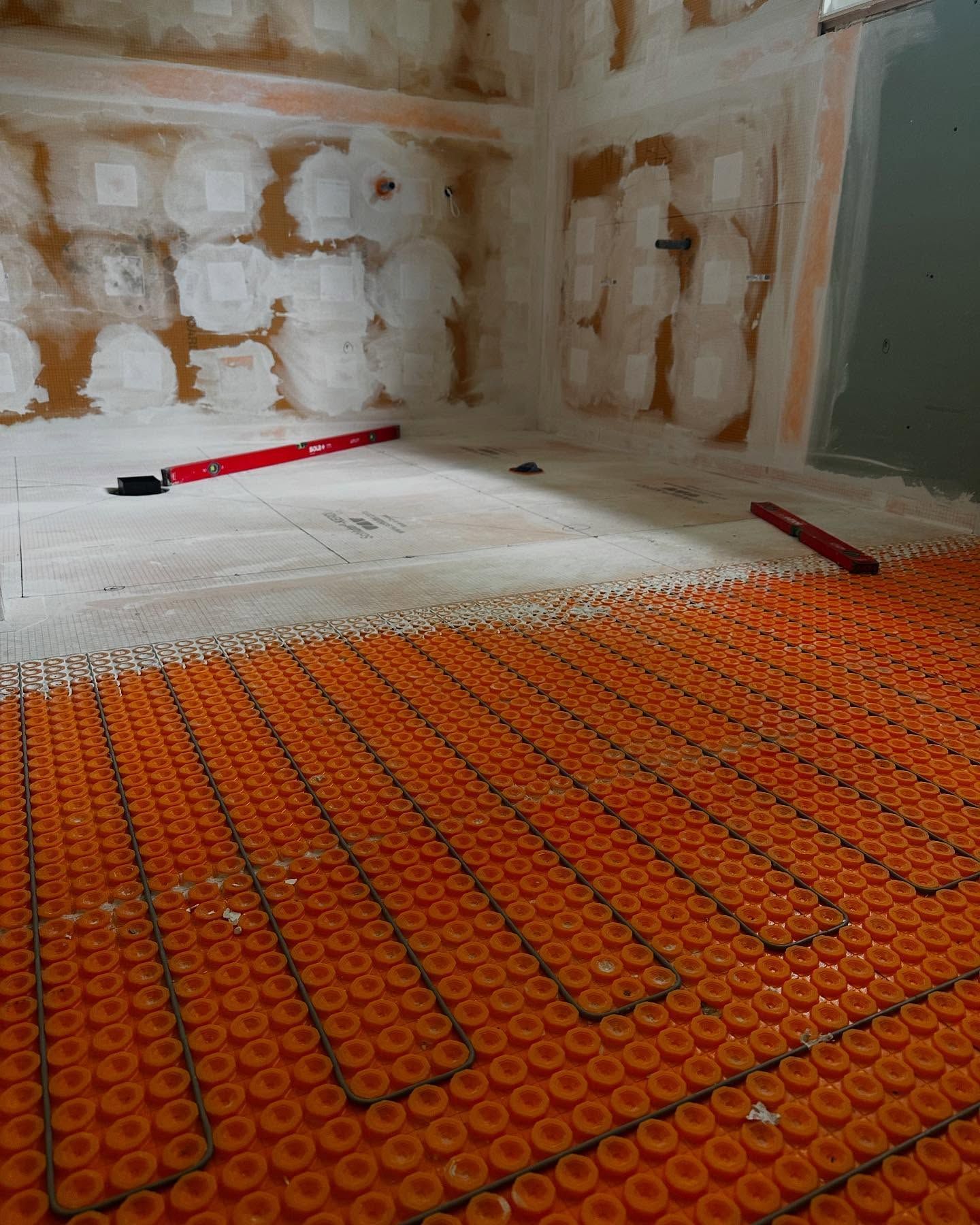 Orange in-floor heating system being installed in a room. Red levels and drywall visible.