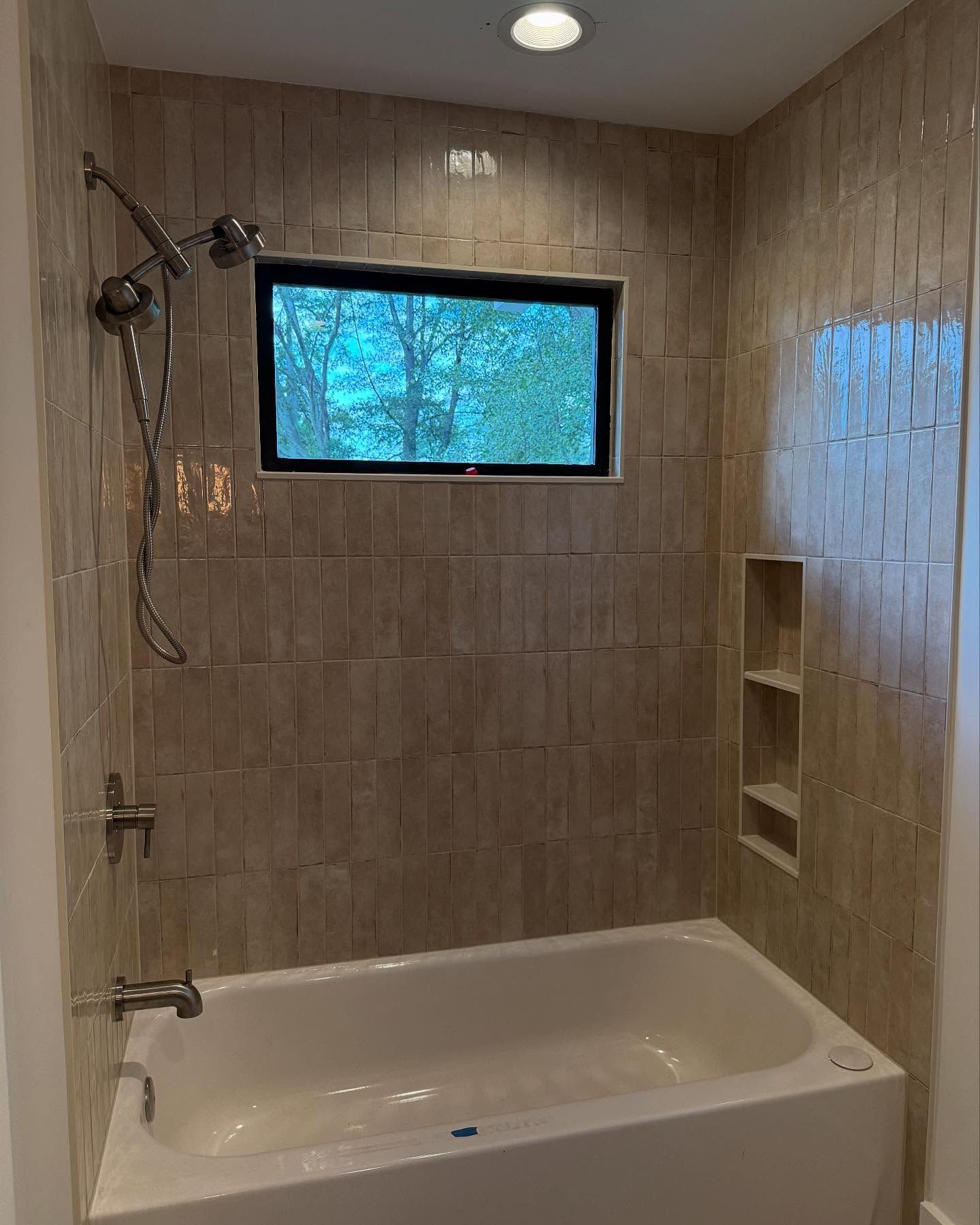 Bathroom with beige tiled walls, white bathtub, window, and silver showerhead.