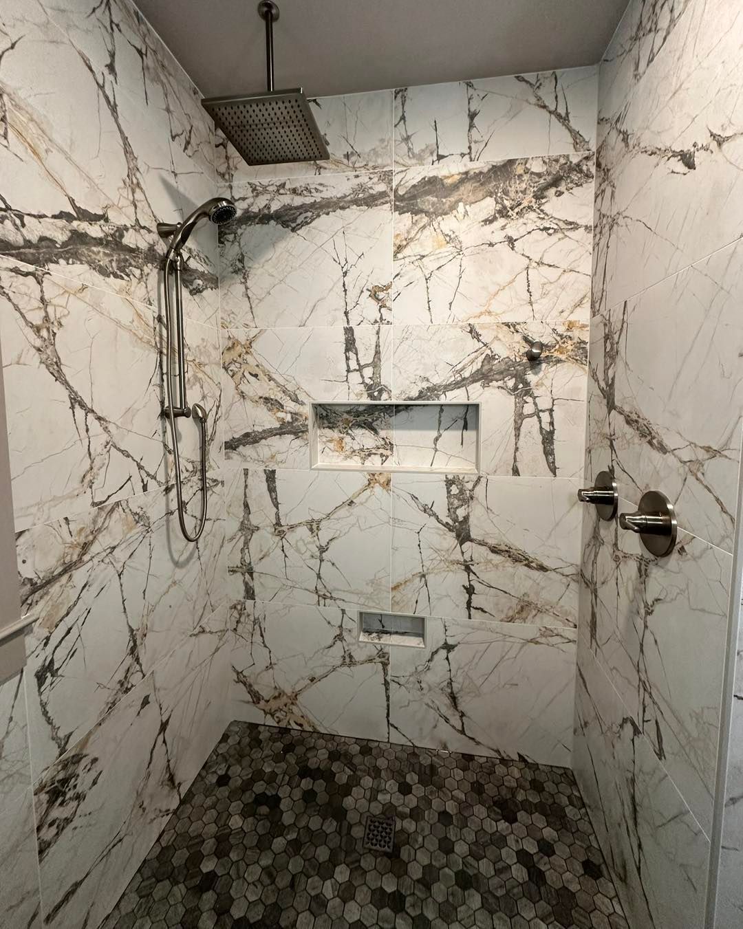Shower with white marble-like tiles, gray grout lines, and a stone-tiled floor. Includes two built-in shelves.