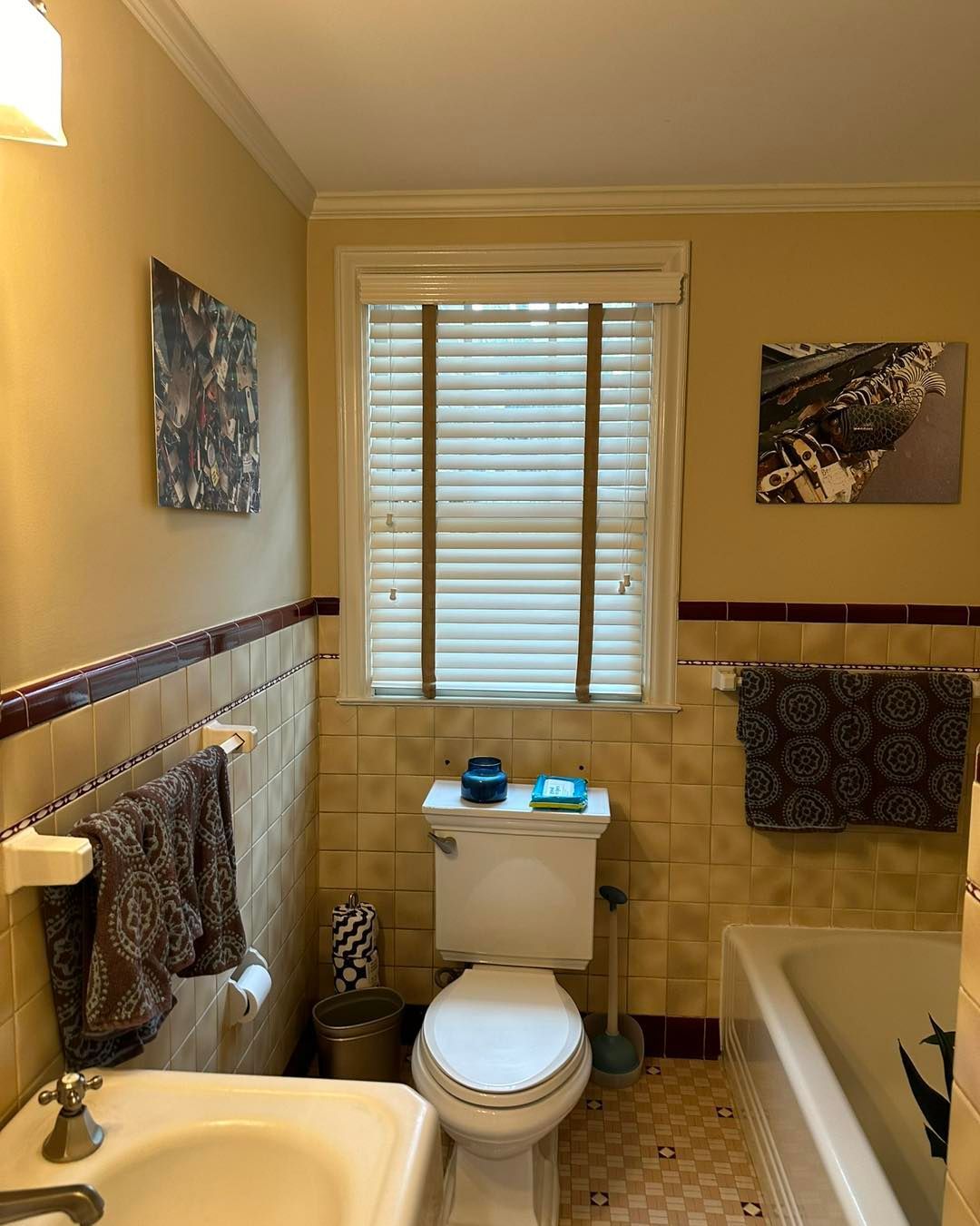 Bathroom with beige walls, window with blinds, toilet, sink, towels, and art.