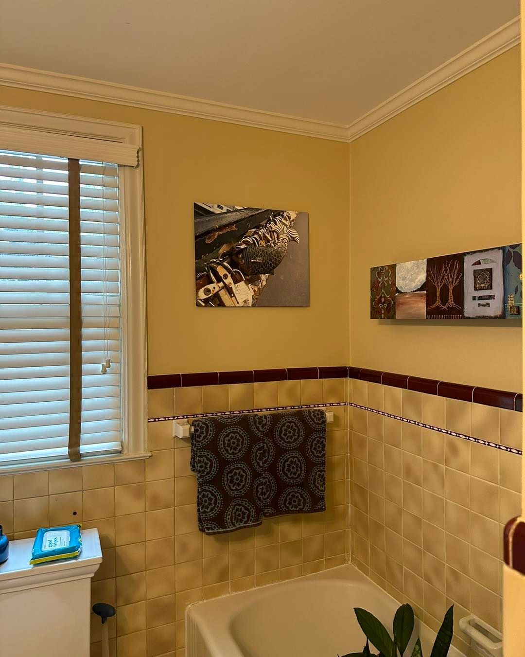 Bathroom with yellow walls, art, towel rack, and a window with blinds.