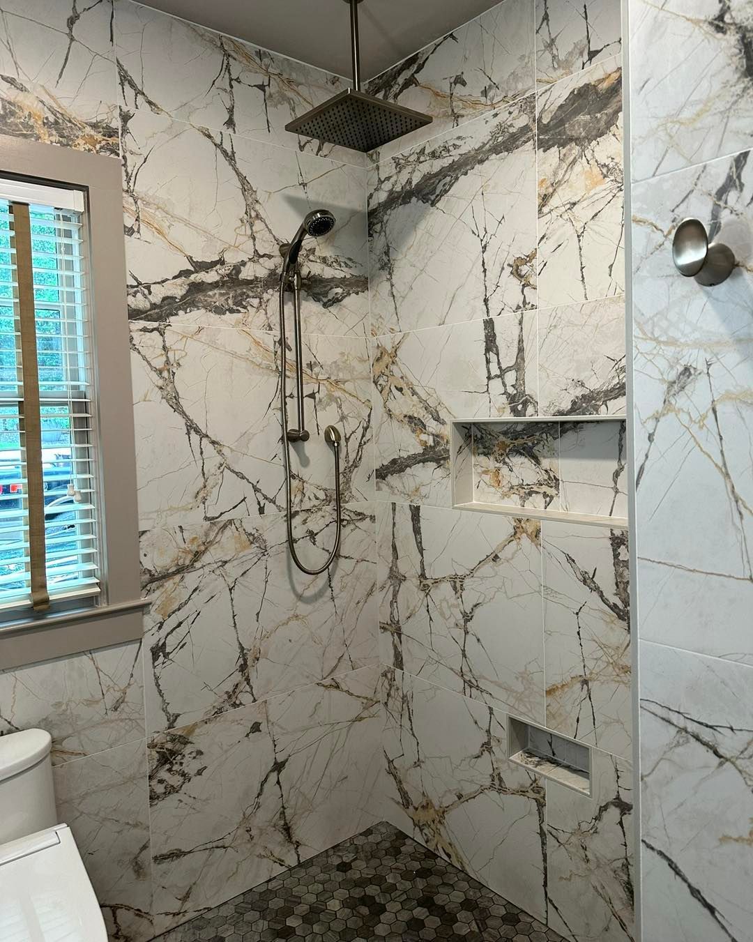 Shower with white marble-look tile, a rainfall shower head, and a built-in shelf; next to a window.