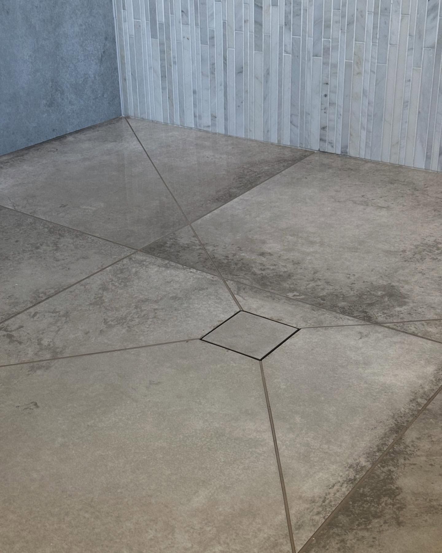 Shower floor with large, light gray tiles angled towards a square drain. Walls are gray and textured.