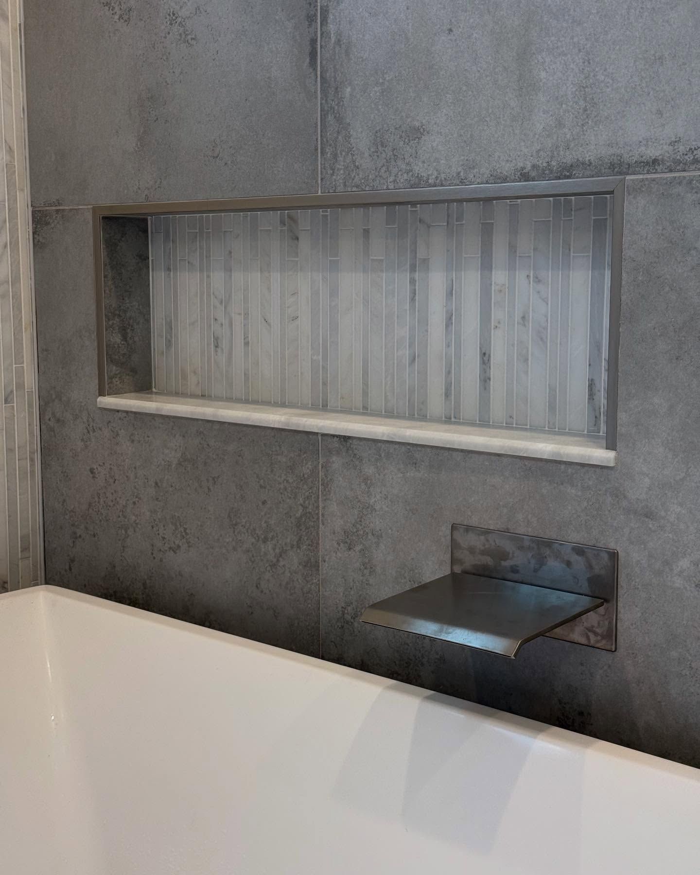 Recessed tile shelf with a metal shelf next to a white bathtub in a gray tiled shower.