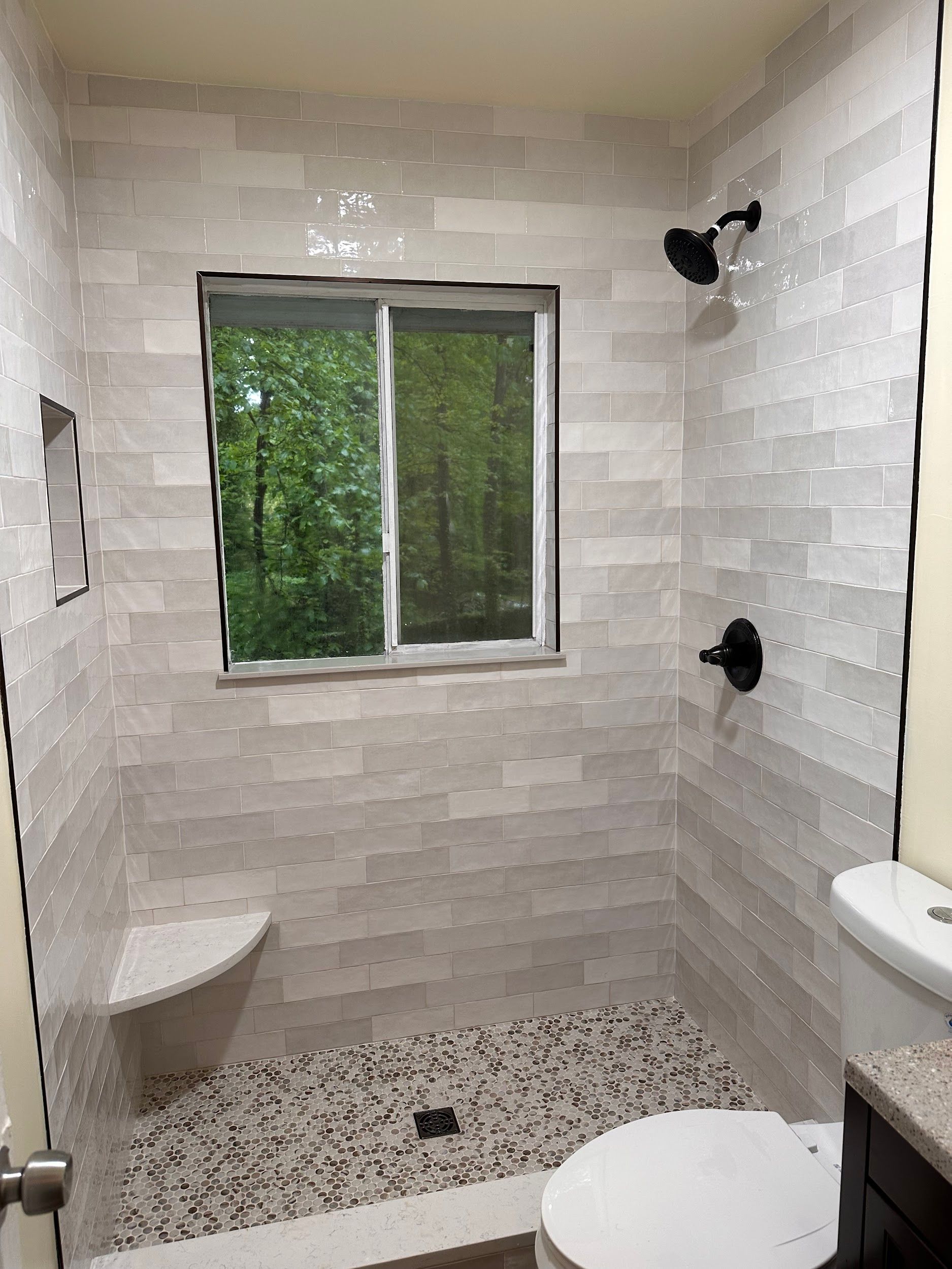 Bathroom with light-colored, textured tile walls, pebble floor, window with green view, and black fixtures.