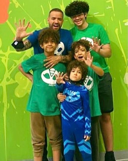Man and four boys waving in front of a green wall. They are wearing matching shirts in shades of blue and green.