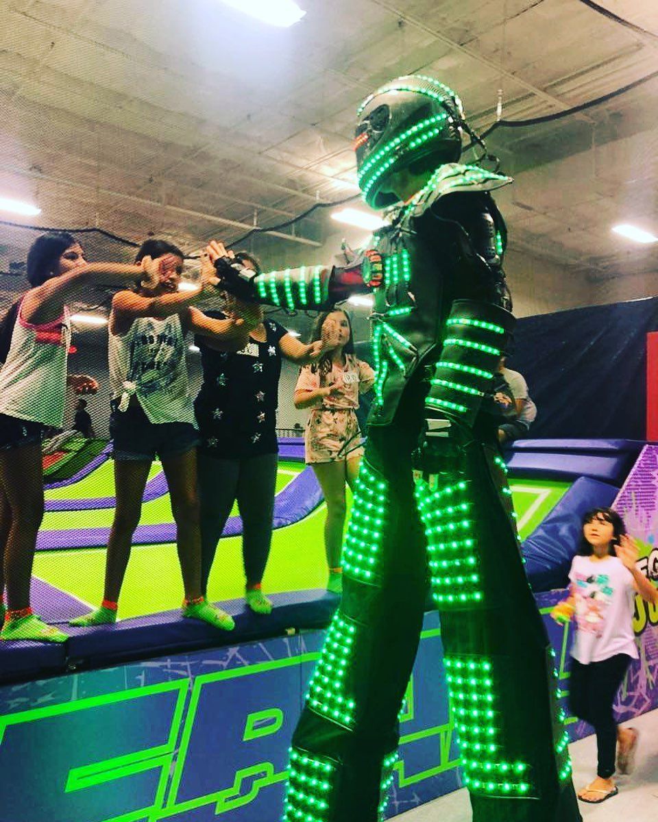 A performer in a green LED suit walks toward a group of people at a trampoline park.