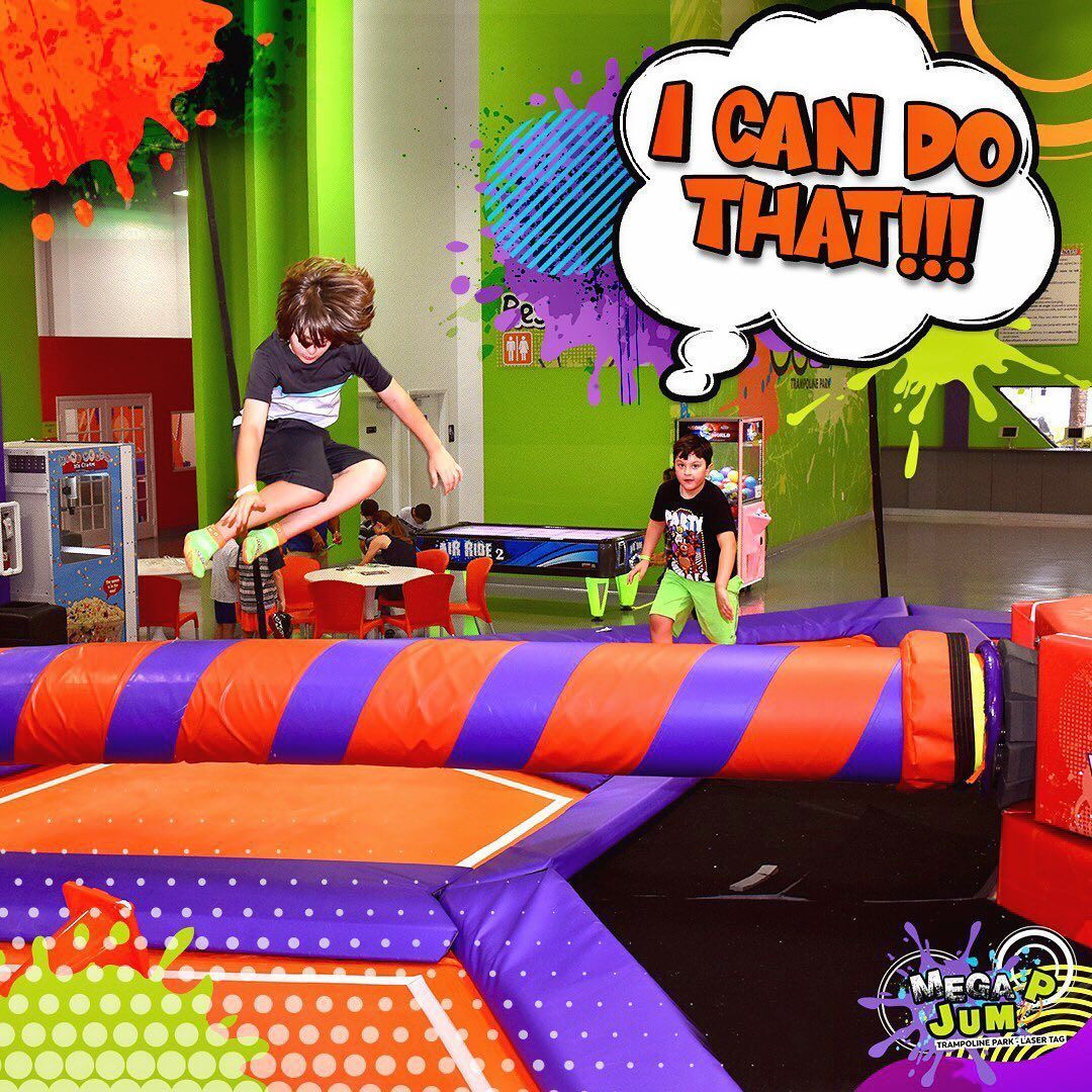 Two boys jumping over a padded obstacle at an indoor recreation center. One boy says