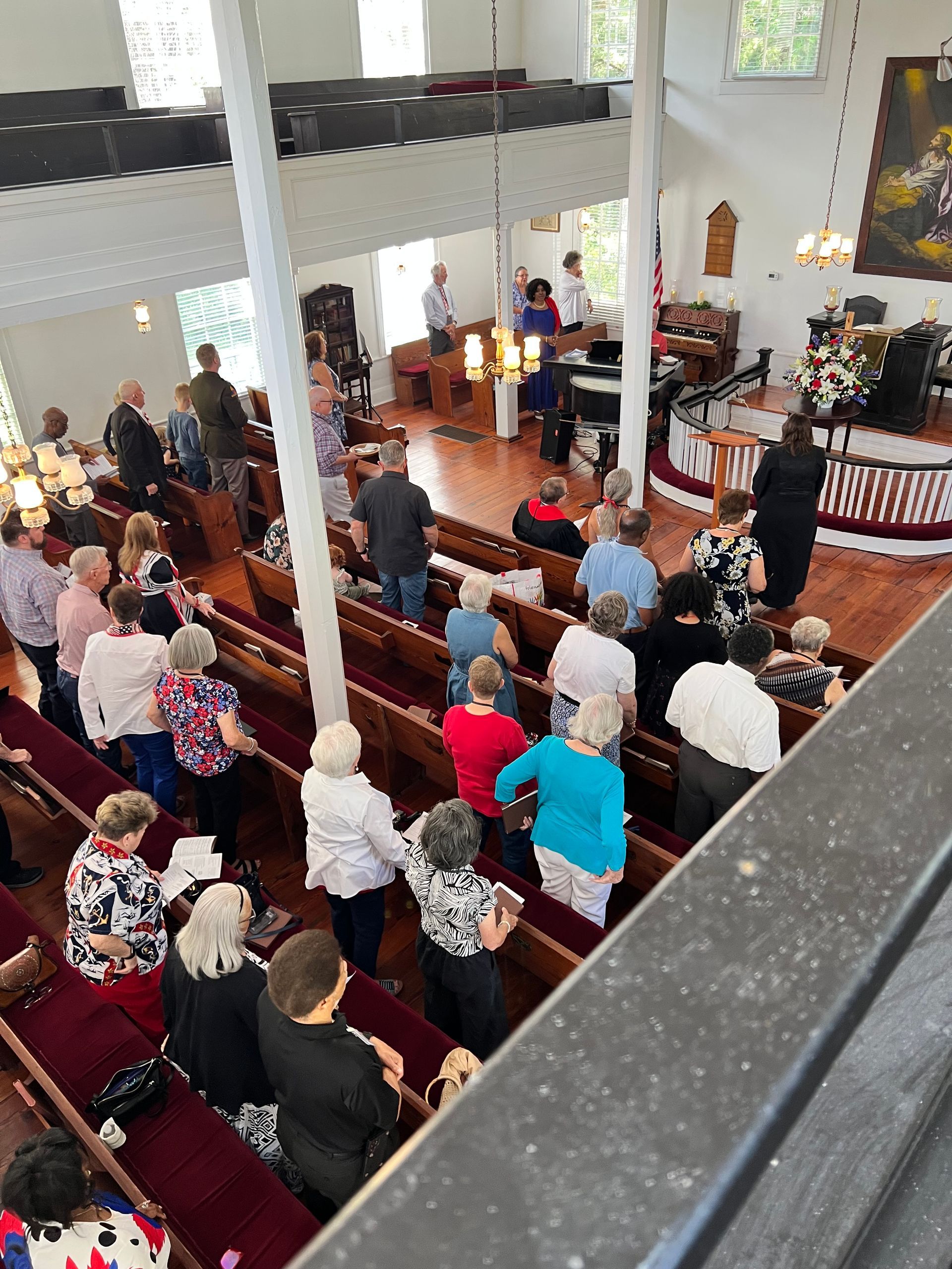 People in a church, some standing, some seated in pews. Wooden interior, with a stage at the front.