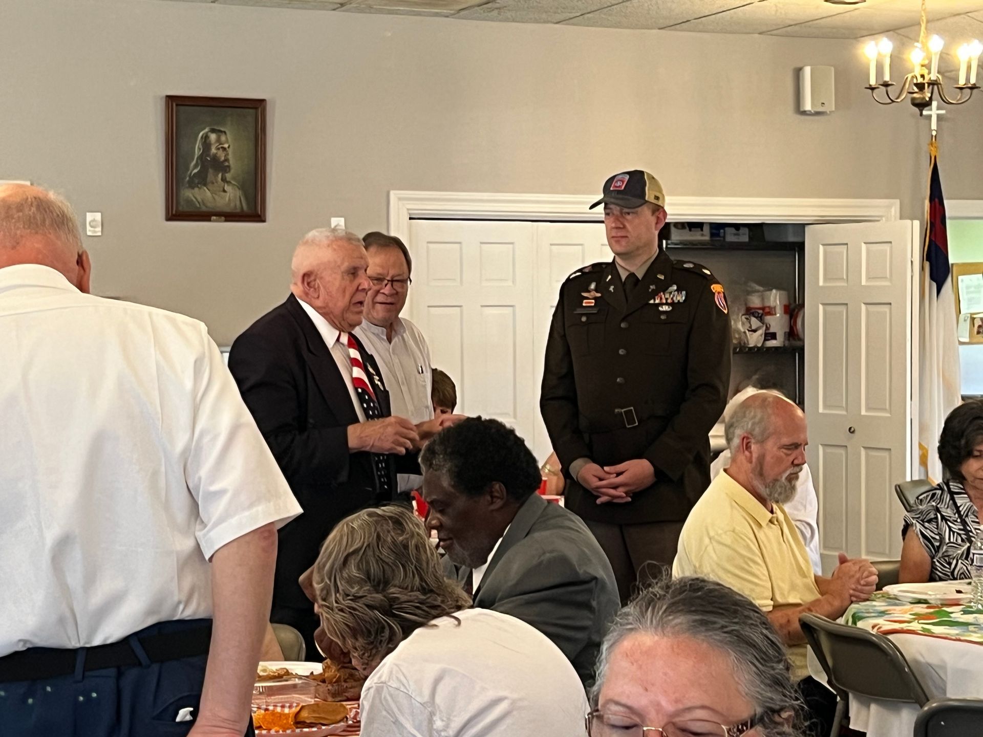Group of people at an event. Man in uniform, others in suits and casual attire, some seated at tables.