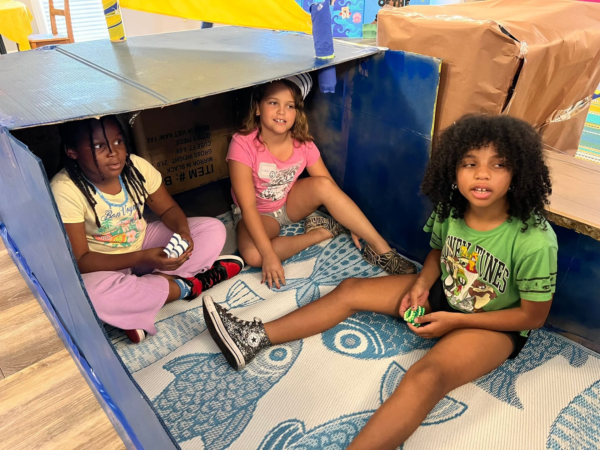 Three girls in a cardboard fort. One in pink, two in green, smiling, sitting on blue fish-patterned mat.