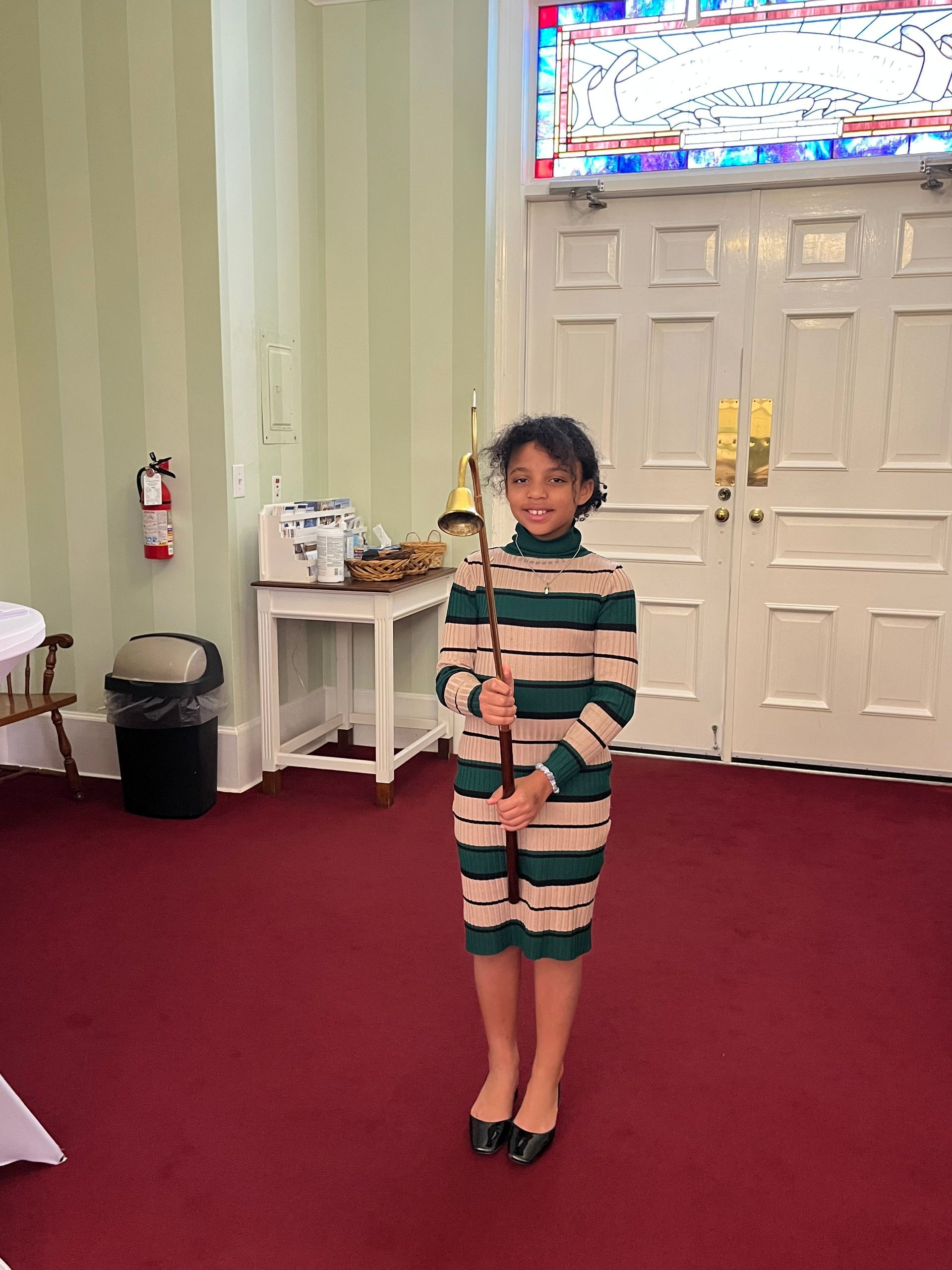 A little girl in a striped dress is holding a stick in a room.